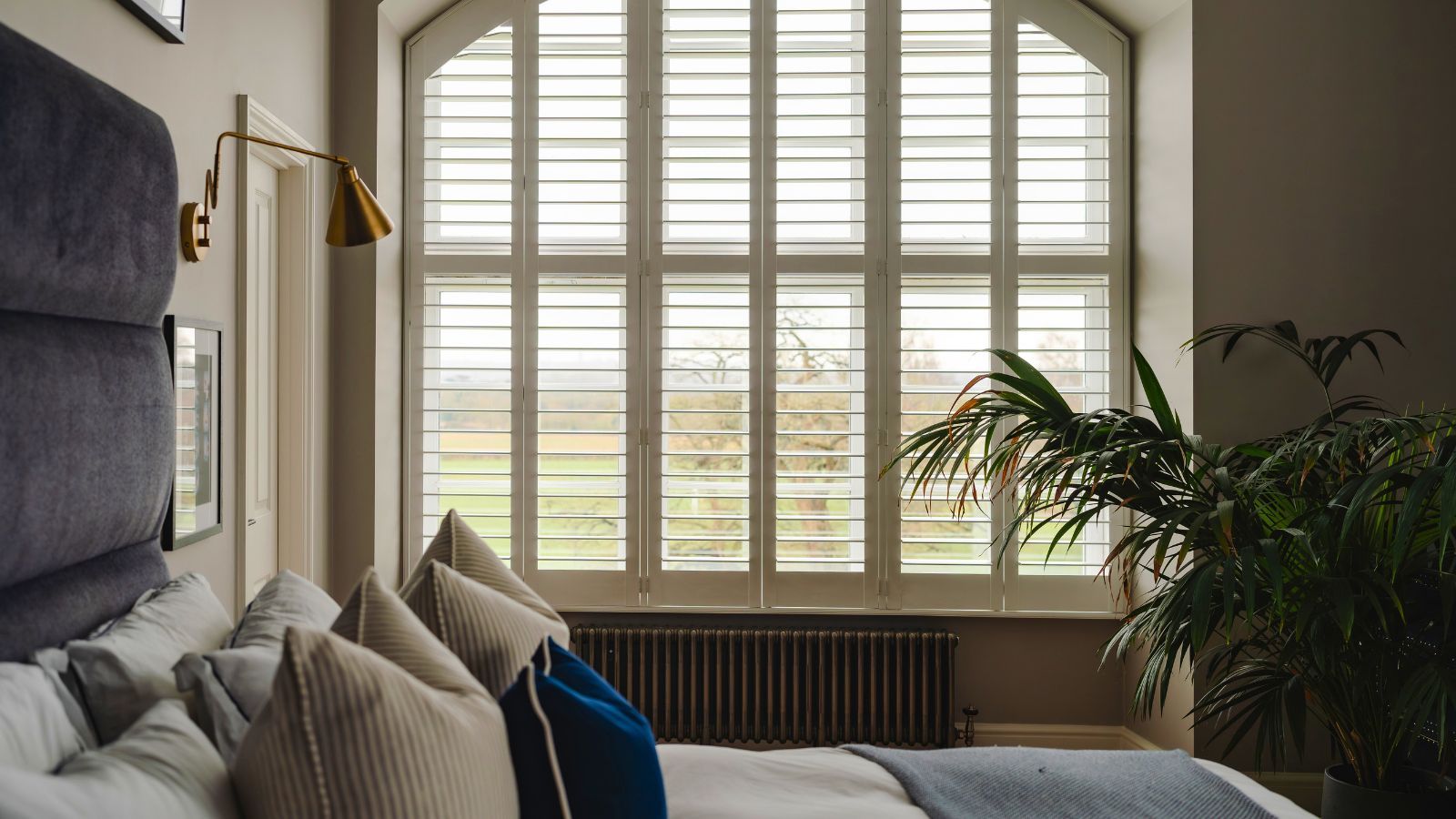 A bedroom with a large arched window and a radiator beneath it. A bed with cream and blue throw pillows on it in the foreground.