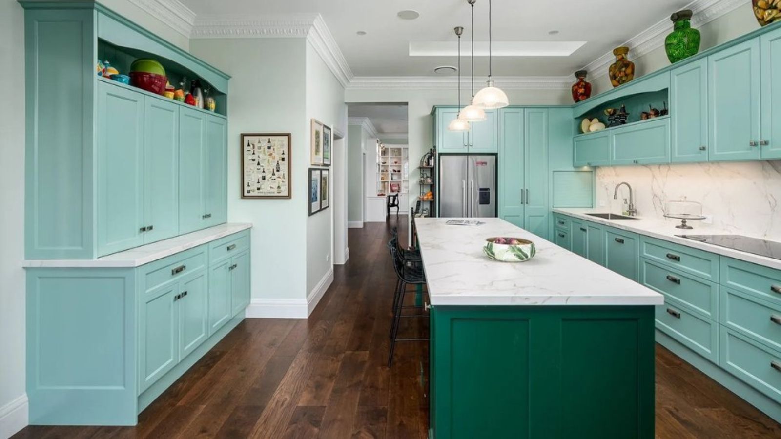 Teal and green kitchen with kitchen island, wooden floors, three hanging lights. Vases and ornaments are positioned on the shelves and above cabinets, and a hallway is pictured in the background.