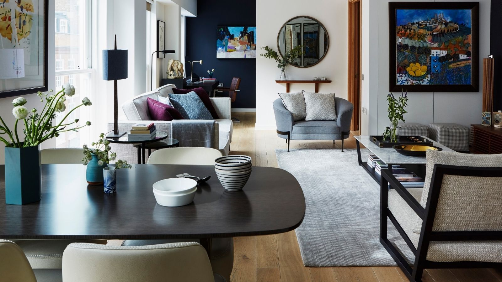 An eclectic open plan living and dining room, with a large black dining table in the foreground, various mismatched sofas and chairs with colorful cushions, and a large grey rug to the right with a large coffee table. The walls are white and dark blue, with mirrors, artwork and shelves hung up, and to the right is a large window that's filling the room with light.