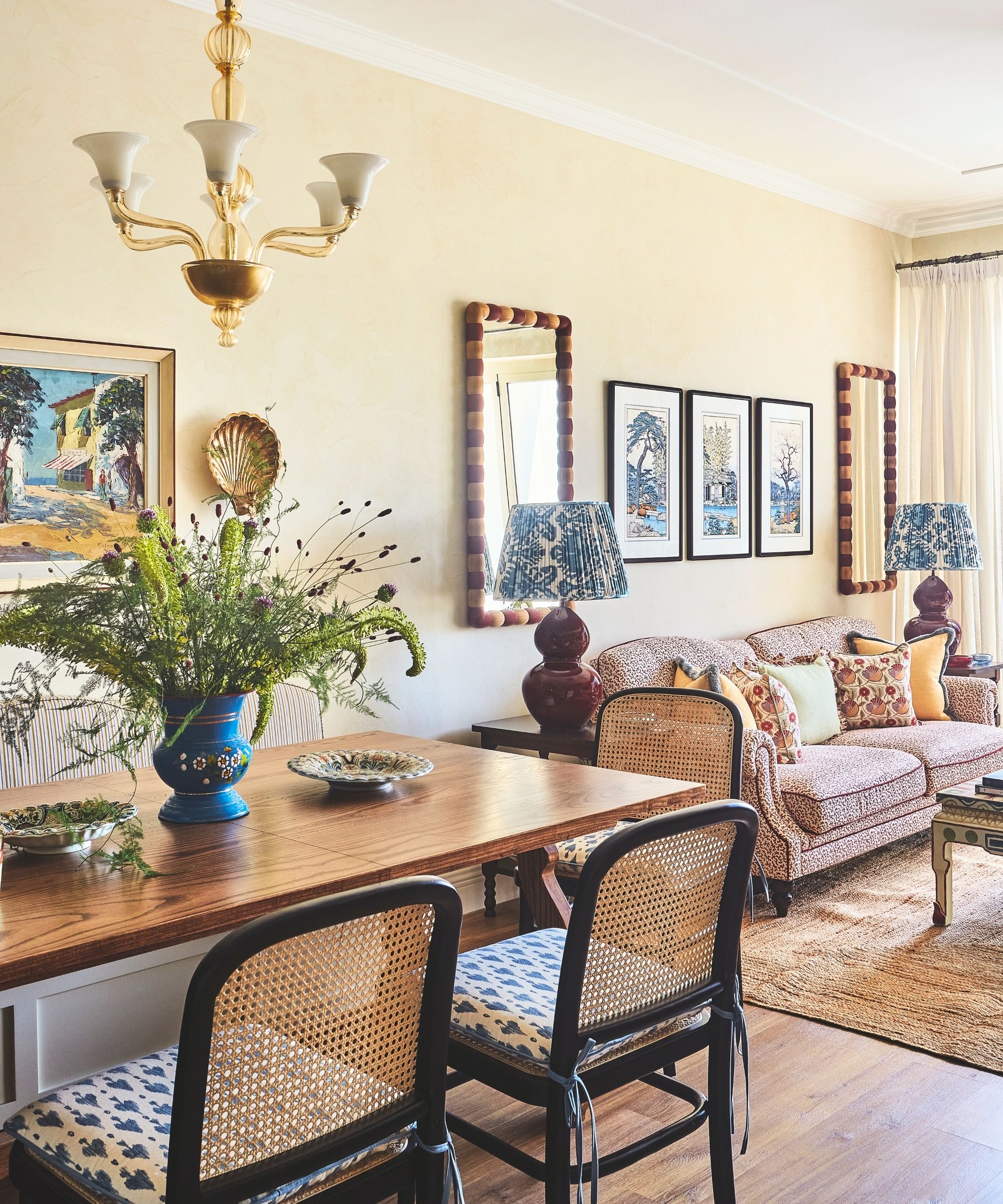 Wohnzimmer mit gemustertem Sofa und Sessel und strukturiertem Seegrasteppich und bunt bemaltem Holzcouchtisch und Kunst an den Wänden dahinter mit symmetrischen Spiegeln