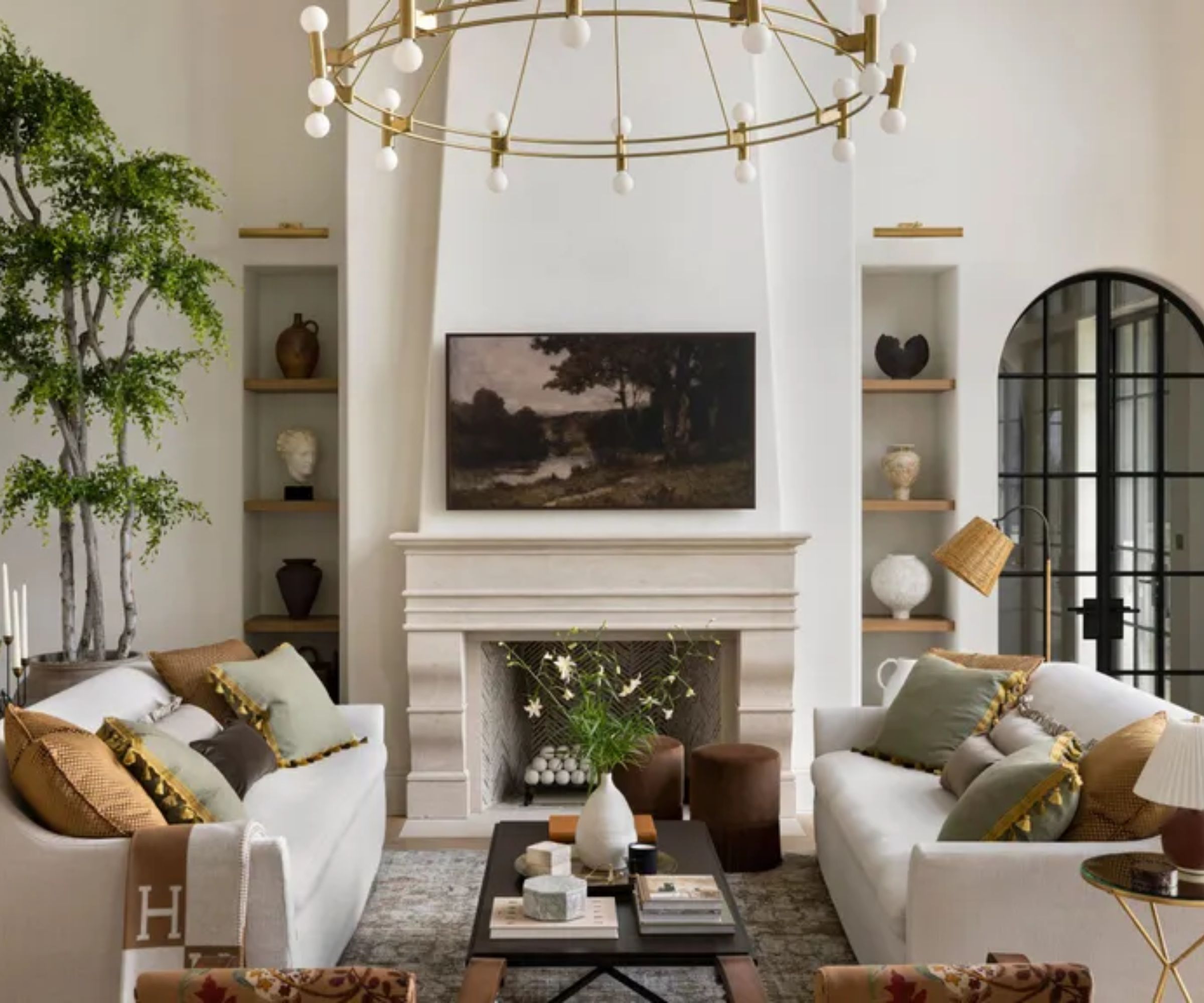 Ein helles Wohnzimmer mit zwei weißen Sofas vor einem Steinkamin. In der Ecke steht ein großer Baum und am Kamin stehen Regale mit Töpferwaren. Darüber hängt ein runder goldener Kronleuchter und hohe, schwarz gerahmte Fenster lassen natürliches Licht herein. In der Mitte steht ein Couchtisch mit Büchern und Blumen.