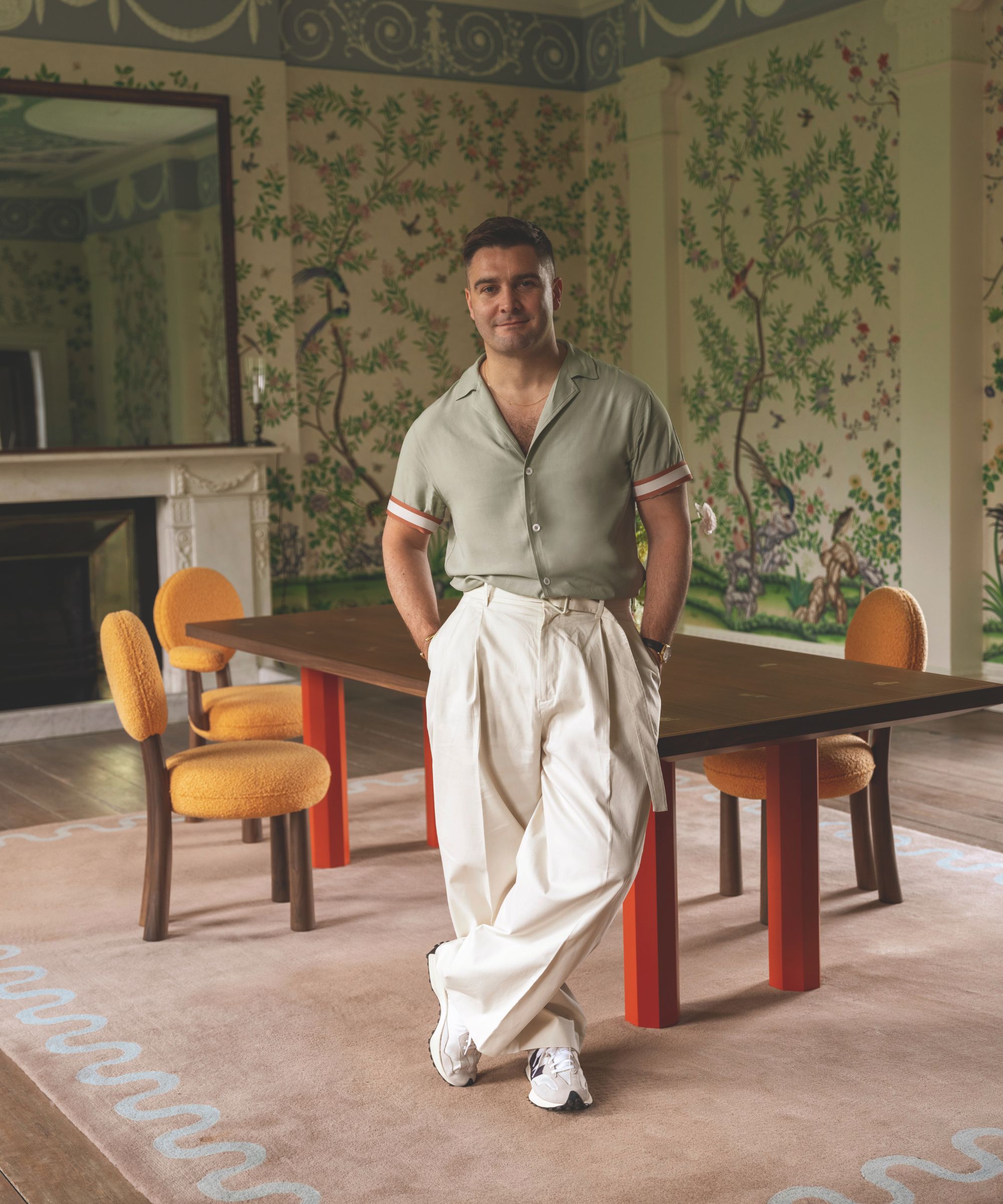 Ein Mann in einem graugrünen Poloshirt und einer cremefarbenen Hose steht in einem Esszimmer mit einem großen rosa Teppich und einem Esstisch mit orangefarbenen Stühlen