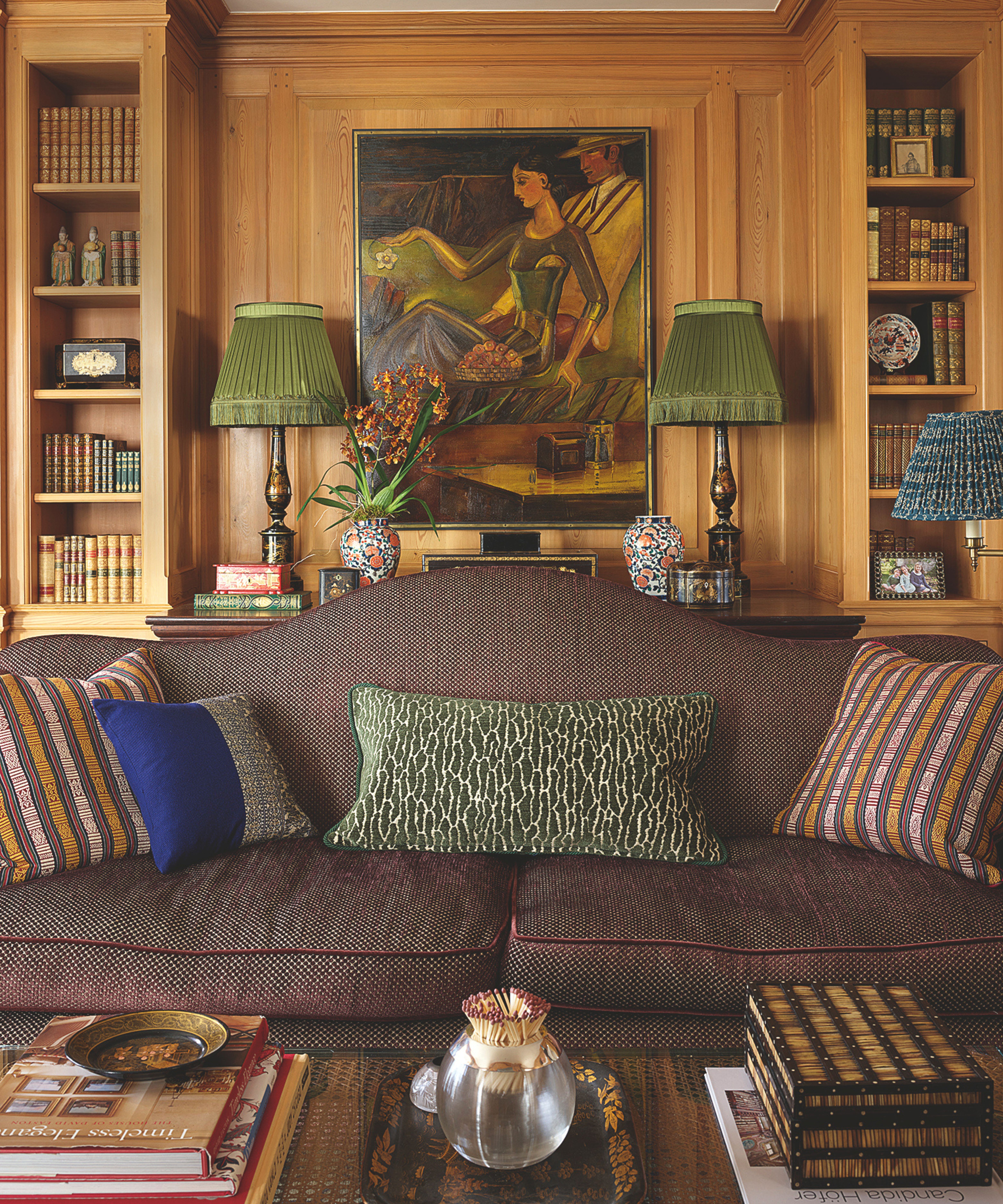 Gemütliches Wohnzimmer mit heller Holzvertäfelung, eingebauten Bücherregalen, einem braun gemusterten Sofa und zwei Lampen mit plissierten grünen Schirmen neben einem Gemälde