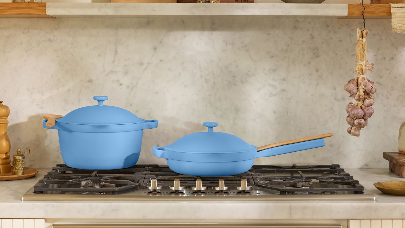 A blue Dutch oven pan, and a matching blue frying pan with lids on a black gas burning stove top. There is a matching greige stone backsplash and countertop, and garlic bulbs hand from a shelve above.