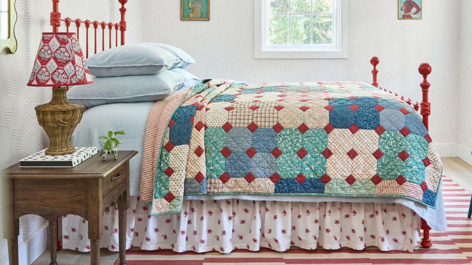 a cottage style bedroom with a rug striped rug, red wrought iron bed, blue linens, a floral bed skirt and patchwork quilt with a wooden nightstand and rattan lamp