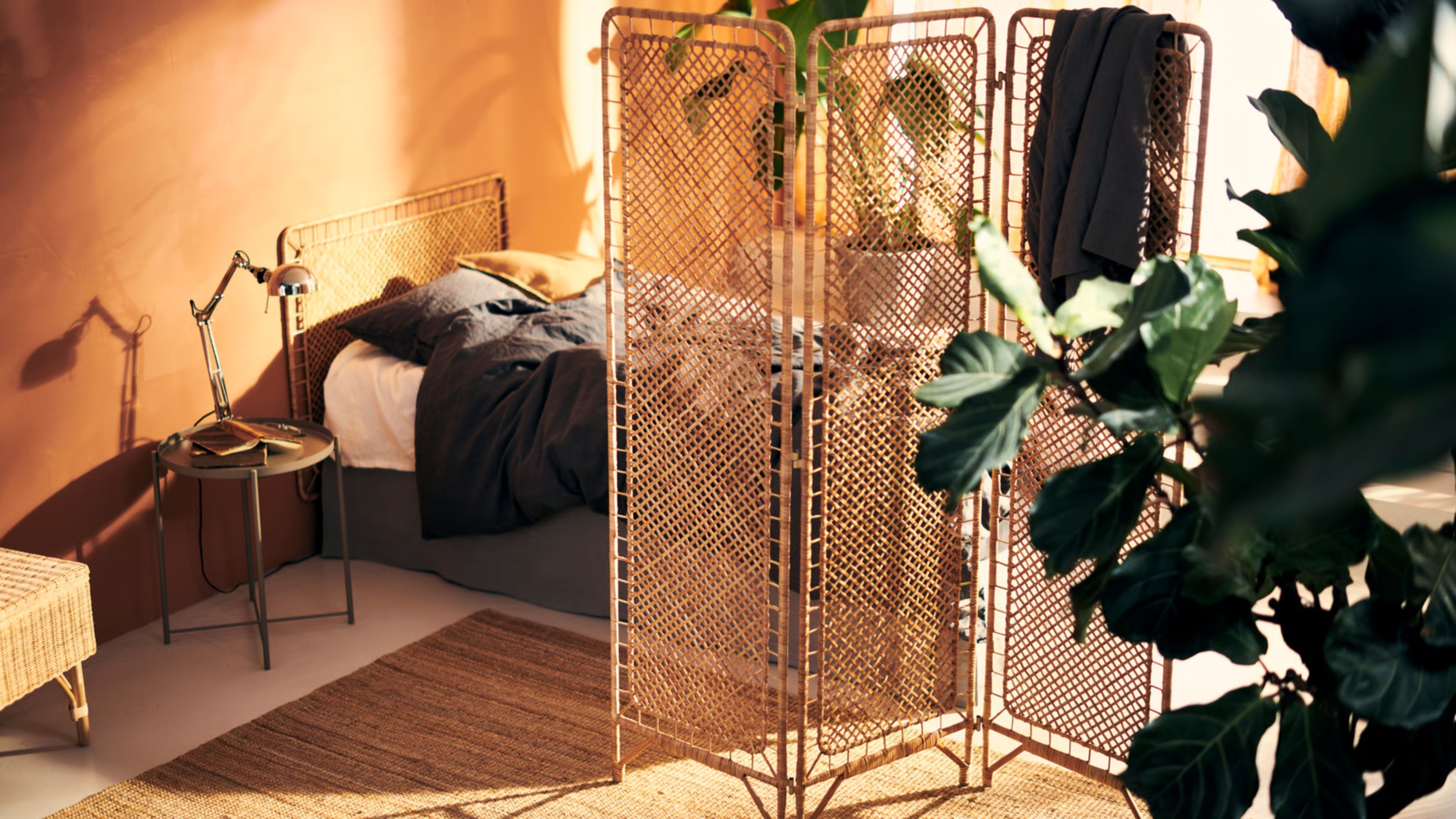 Ein orange gestrichenes Schlafzimmer mit einem niedrigen Bett mit grauer Bettwäsche und einem Kopfteil aus Rattan. Ein Raumteiler aus Rattan trennt den Raum