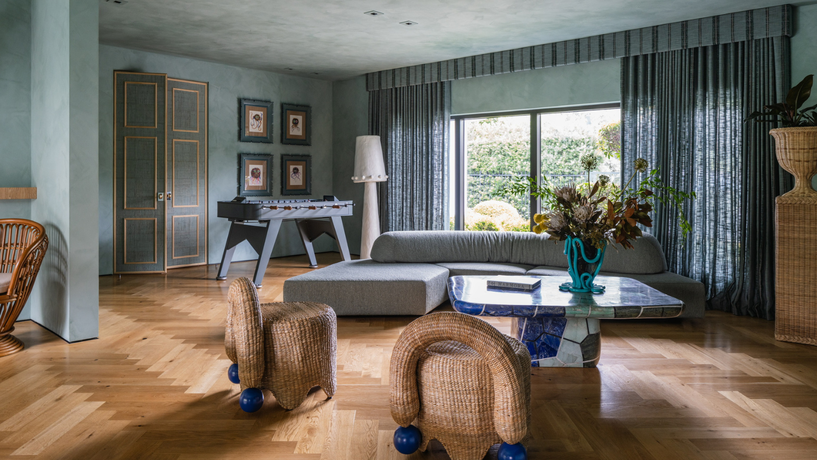 ein großes Wohnzimmer mit hellblau gestrichenen Wänden und Decken, blauen Vorhängen, die die großen offenen Fenster versperren, einer blauen Couch und Korbstühlen 