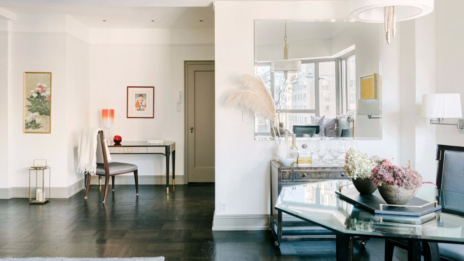 Bright living space with glass tables, chairs, white walls, framed art, a large mirror, and a wooden floor.