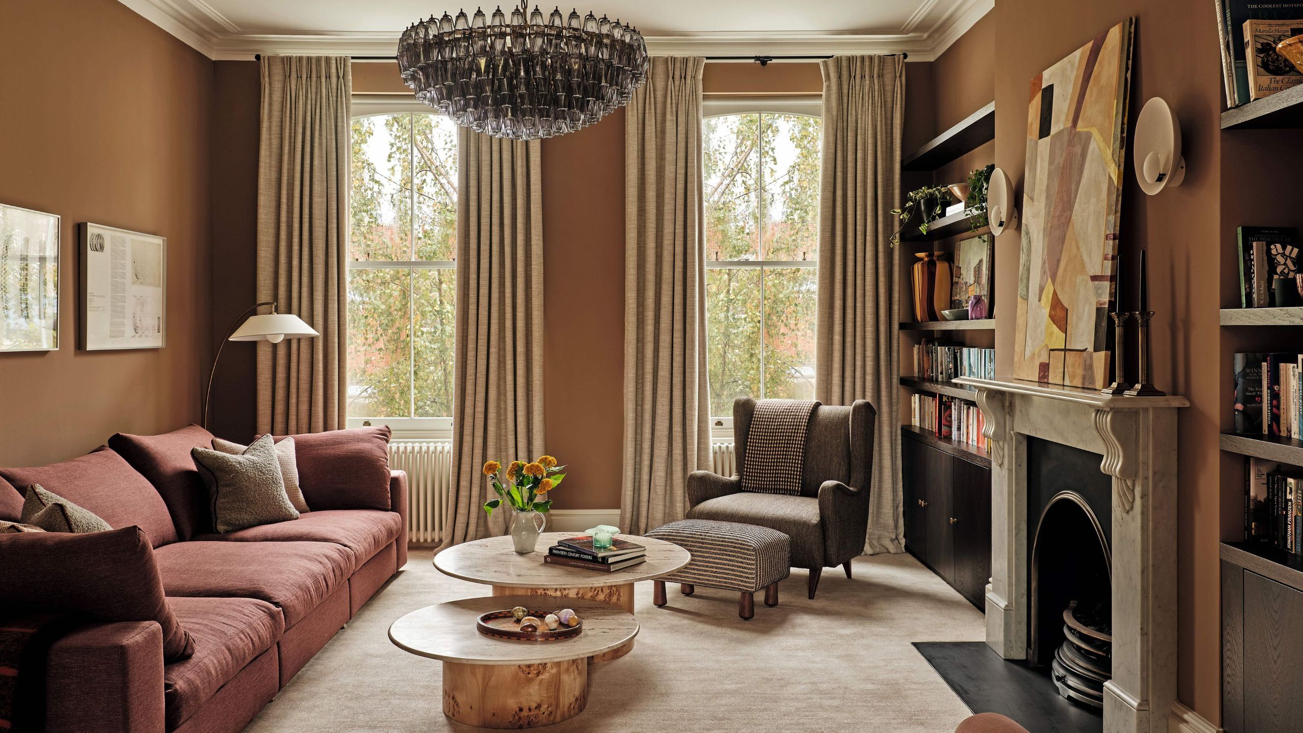 A warm, earth-toned living room featuring terracotta walls, a dusty rose sectional sofa, and a large tiered glass chandelier. Two organic-shaped wooden coffee tables sit on a plush beige rug before a marble fireplace.