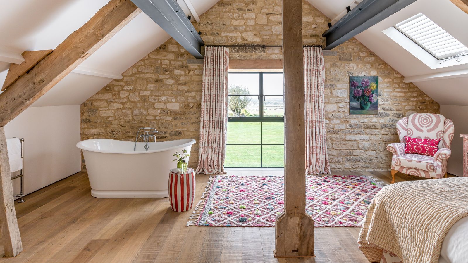 Ein Schlafzimmer im Dachgeschoss mit Holzboden, freiliegenden Balken und einer freiliegenden Ziegelwand. Rechts im Zimmer steht ein Bett mit einer blass pfirsichfarbenen Decke und links eine große weiße Badewanne. Außerdem verläuft in der Mitte des Raumes ein großer Holzbalken, dahinter ein gemusterter rosa-weißer Teppich und links davon ein gemusterter rosa-weißer Polstersessel.