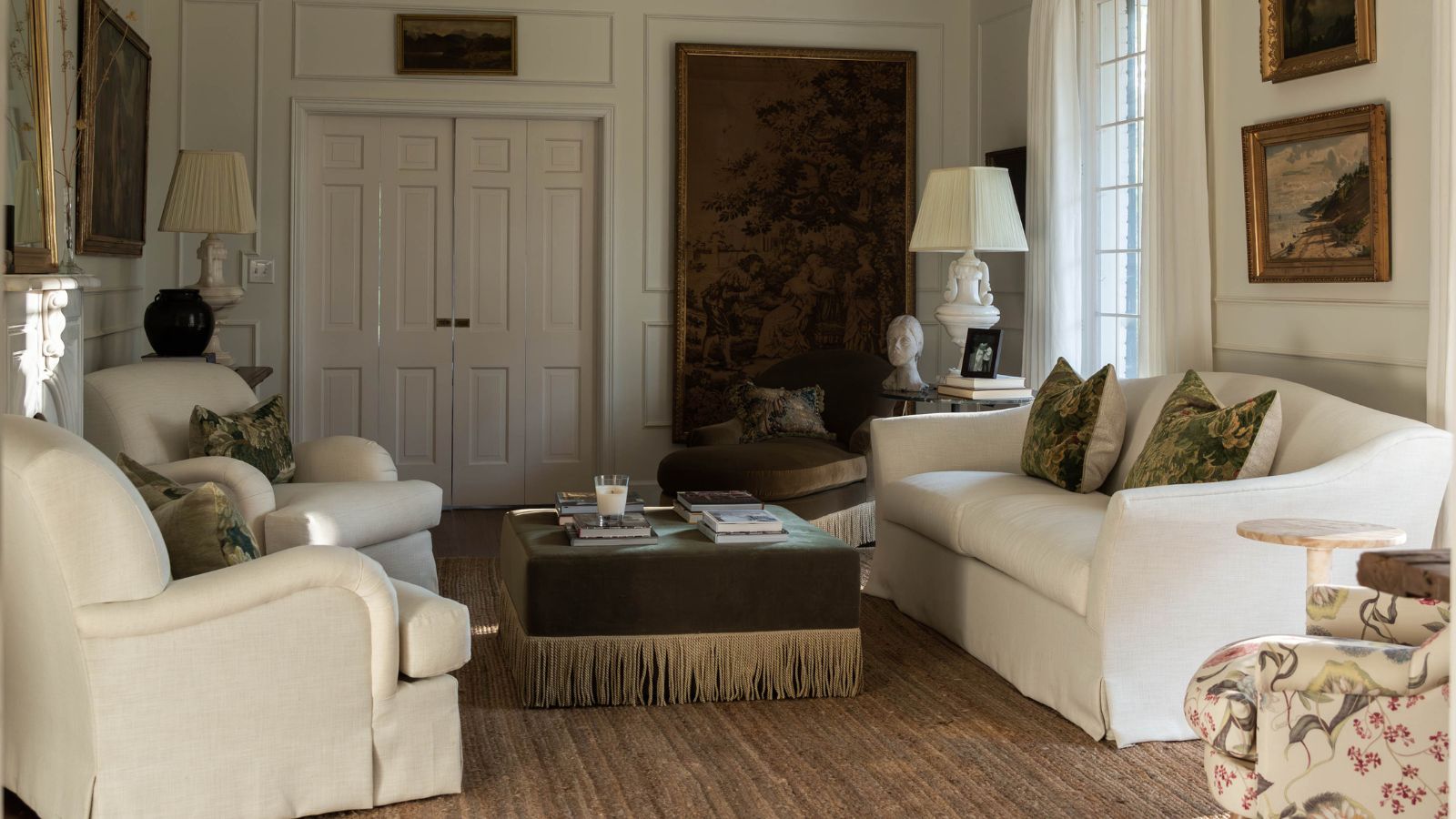 Ein traditionelles weißes Wohnzimmer mit einem großen antiken Wandteppich und Vintage-Kunstwerken, Wandleisten, zwei weißen Sesseln, einer Leinencouch und einem Ottomane mit grünen Samtfransen