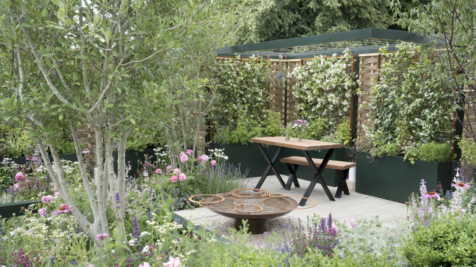 Ein moderner Garten mit Kletterpflanzen, Bäumen, Blumenrabatten, einem Wasserspiel sowie Terrassentischen und -stühlen bei der Hampton Court Flower Show