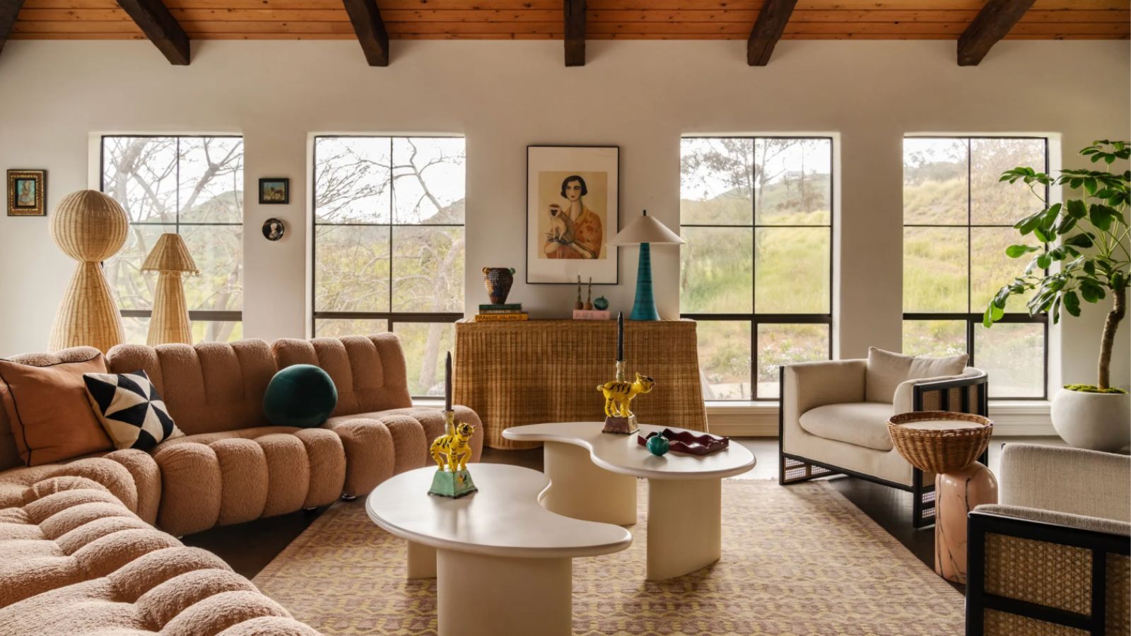 Large living room with plump brown sofas, abstract tables, patterned rug, armchair, large shelf, ceiling with wooden beams, and four large windows revealing outdoor space.