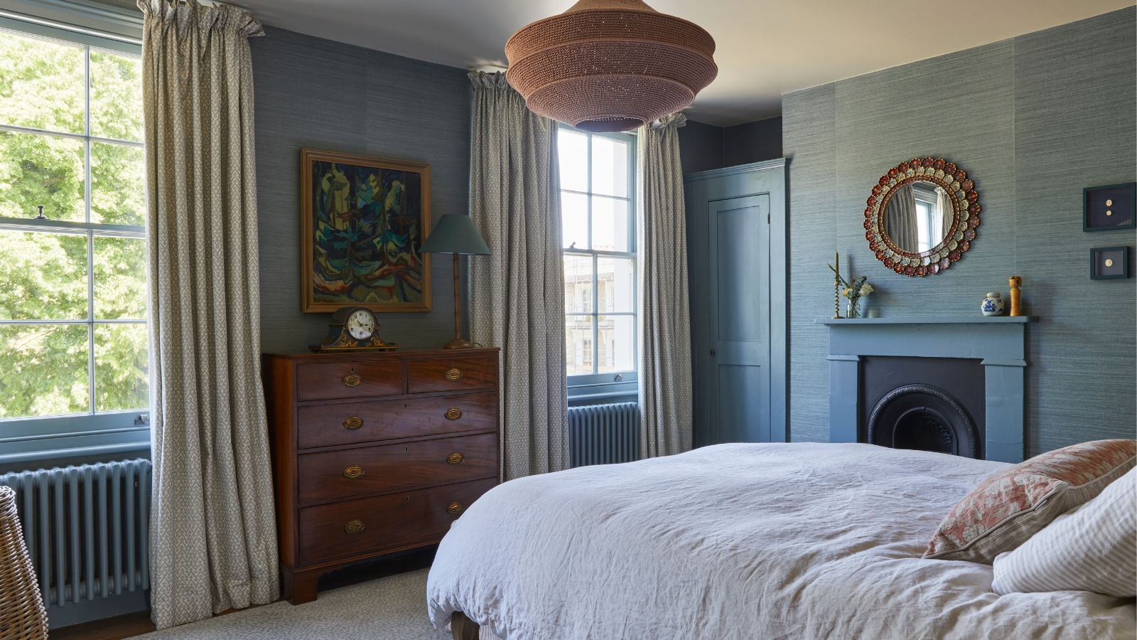 Bedroom with blue grasscloth walls, mahogany dresser, abstract painting, rattan pendant light and large bed