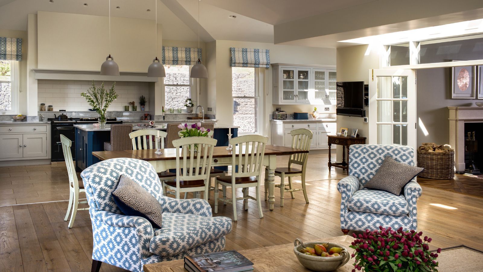 A cozy kitchen space with a large dining table and seating area with patterned blue and white upholstered chairs. The cabinets are white, with a tiled backsplash and large black AGA oven, and the flooring is wood.