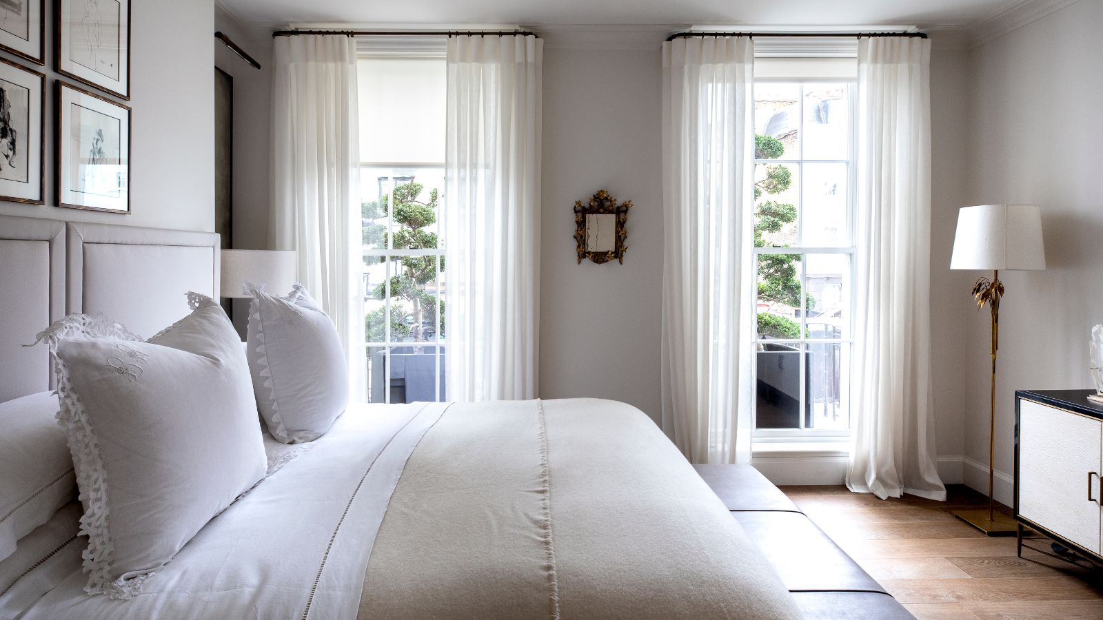 Ein weißes Schlafzimmer mit einem Doppelbett mit weißer Bettwäsche, weißen Wänden, Holzböden, einer goldenen Lampe mit weißem Lampenschirm, schwarz-weißen Schränken und zwei großen Fenstern mit transparenten weißen Vorhängen.