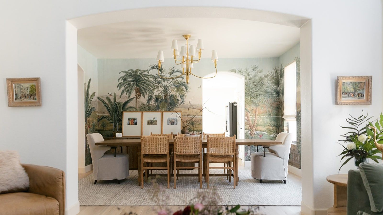White interior archway revealing dining room with floral scene wallpaper and dining table with rattan chairs on white rug. Pictured in the foreground are an armchair, two framed pictures, and a side table with flowers.