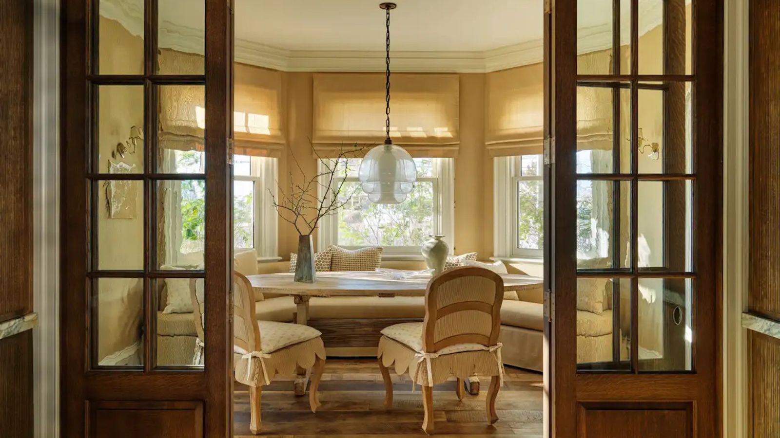 Gemütliche Frühstücksecke in warmem Beige in einem Erkerfenster mit antiken französischen Holztüren, gelben Jalousien und zwei Esszimmerstühlen mit gewellten Polstersitzen