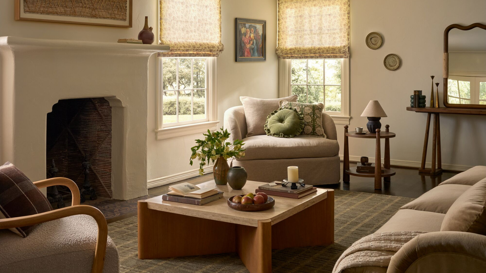 ein Wohnzimmer mit einem großen Kamin, einem quadratischen Couchtisch, einem Akzentstuhl mit Kissen darauf, einem kleinen Beistelltisch und Stoffjalousien vor den Fenstern in der Ecke