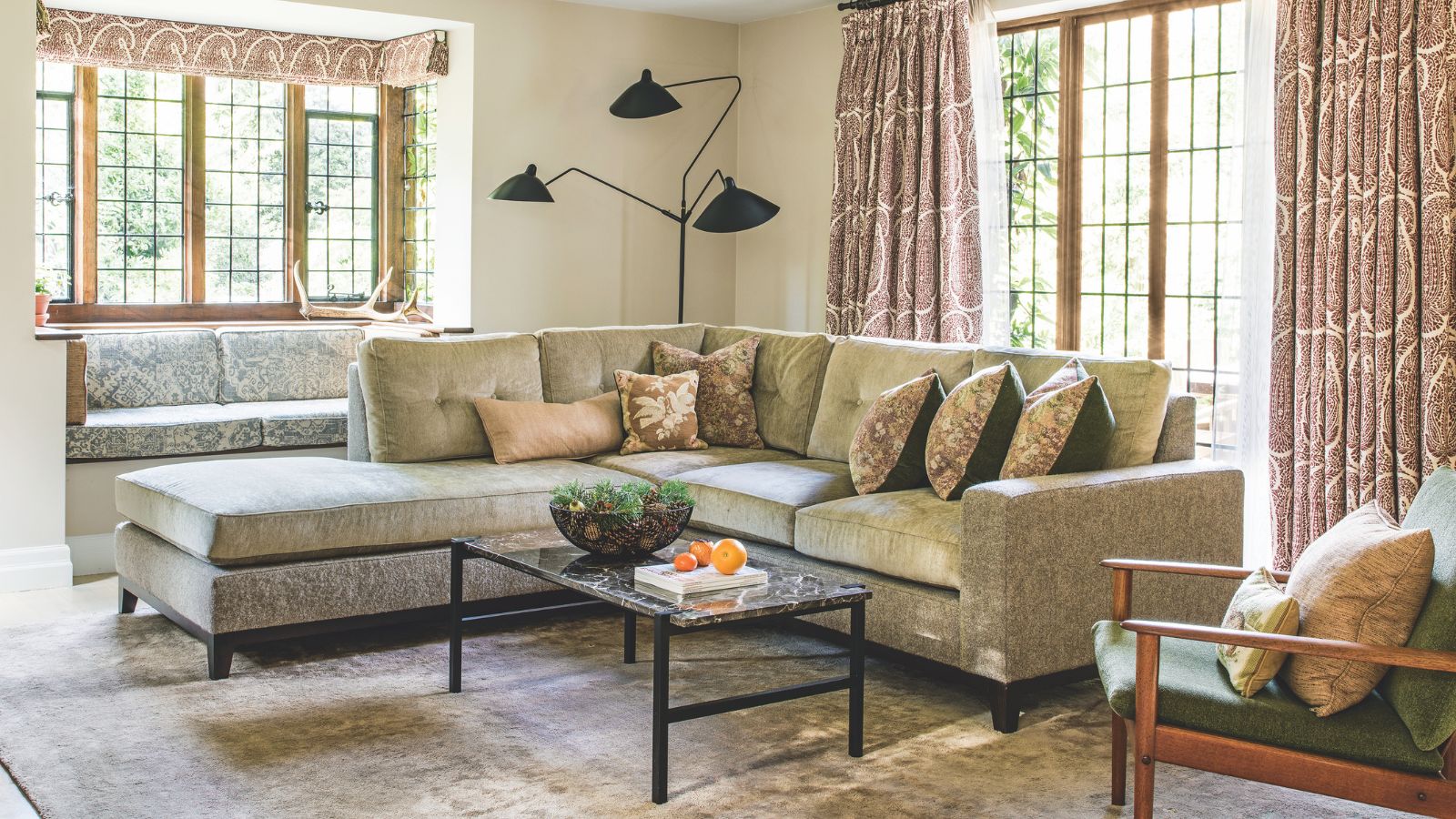 Ein gemütliches Wohnzimmer mit einer großen L-förmigen Samtcouch mit Kissen, einem großen Samtteppich, einem Marmor-Couchtisch und großen Fenstern mit gemusterten Vorhängen. In der hinteren Ecke steht eine große schwarze Lampe und im Vordergrund ein grüner Stuhl mit Holzgestell und zwei Kissen.