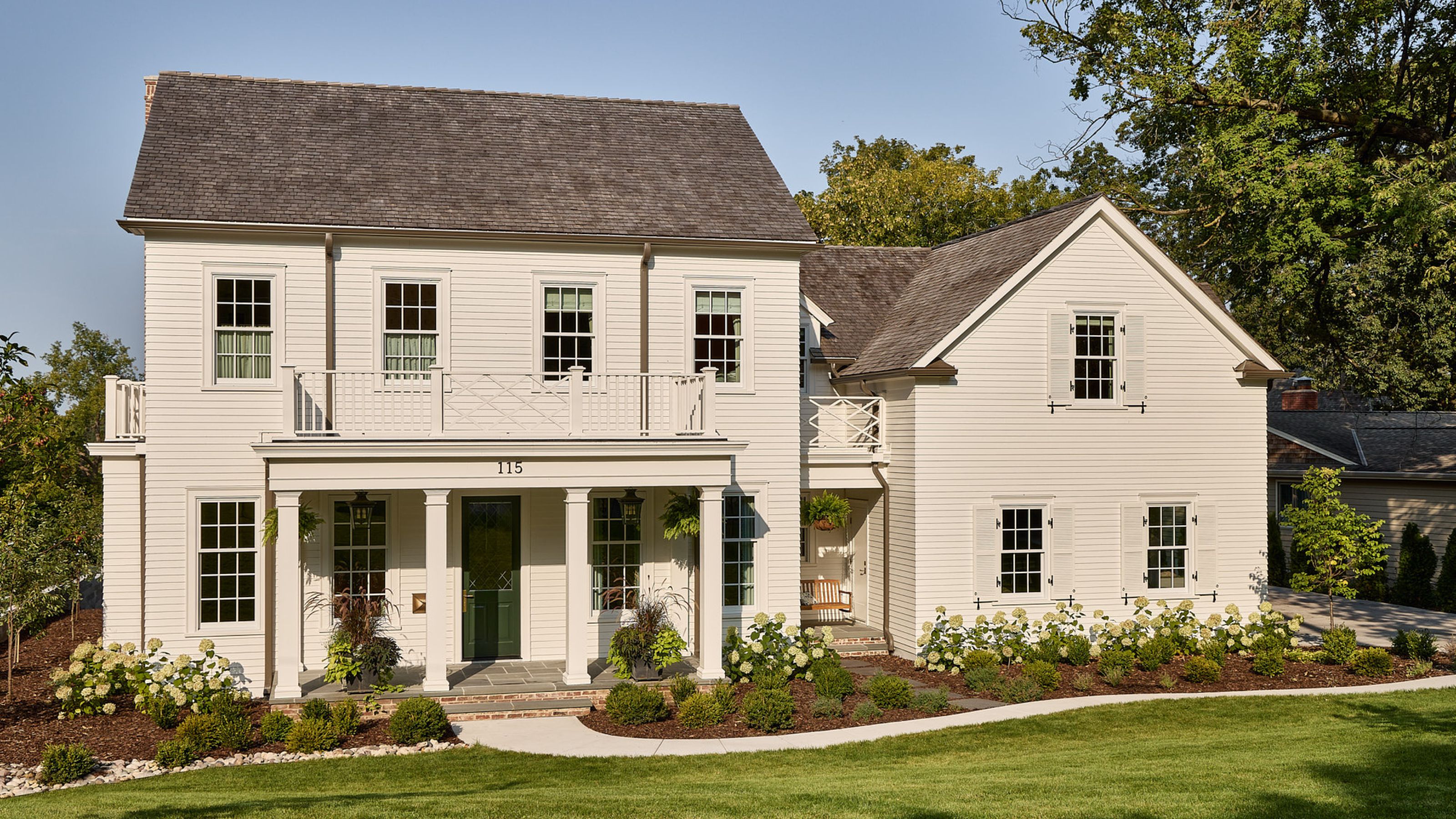 Ein großes Haus von außen mit warmweißen, holzverkleideten Wänden, einer Veranda mit vier Säulen, Fenstern im traditionellen Stil und einer dunkelgrünen Haustür. Ein Garten vor dem Haus mit üppigem grünem Gras und einem geschwungenen Weg. 