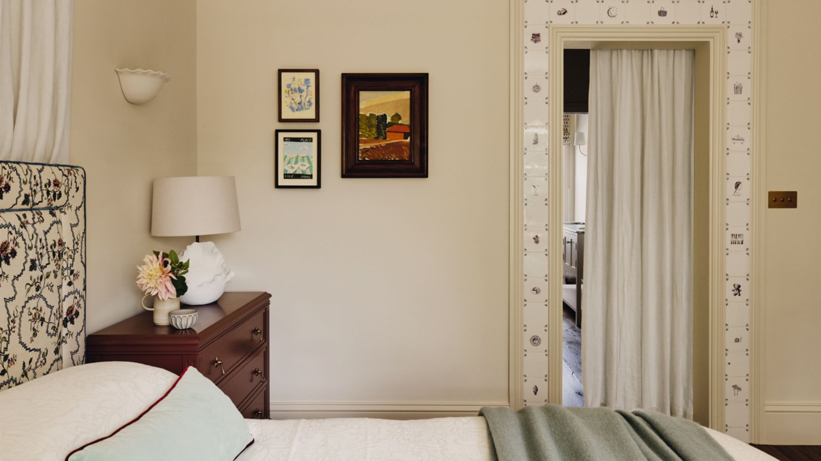 neutrales Schlafzimmer in Farrow & Ball's Dimity mit Türverkleidung aus Delfter Fliesen, kleinem Vintage-Kunstwerk, einem gepolsterten Kopfteil mit Blumenmuster und einem burgunderfarbenen Nachttisch