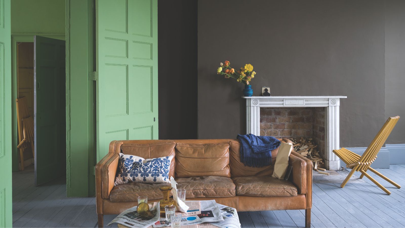 Living room with wooden floorboards, central wall painted dark brown around the large fireplace, a leather couch at the centre of the room, with a lime green painted door and hallway