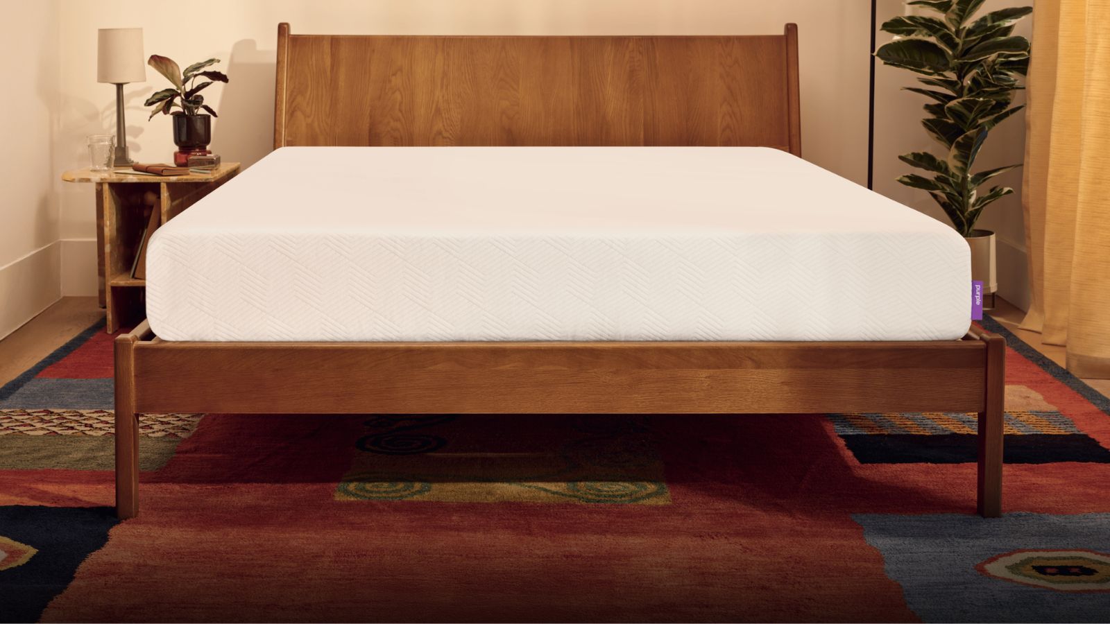 Eine weiße PurpleFlex®-Matratze auf einem Holzbettrahmen mit einem soliden Kopfteil. Auf beiden Seiten des Bettes stehen Zimmerpflanzen, hinter dem Bett befindet sich eine cremefarbene Wand und der Boden ist ein heller, gemusterter Teppich. Rechts ist ein Vorhang, durch den warmes Licht einfällt.