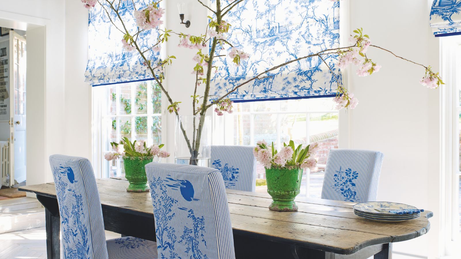 Ein frühlingshaftes Esszimmer mit einem großen Esstisch aus Holz in der Mitte, mit rosa Blumen in grünen Vasen und einem großen Zweig in der Mitte des Tisches. Rund um den Tisch stehen vier gepolsterte Stühle mit einem blau-weißen Toile de Jouy-Muster, dahinter befinden sich große Fenster mit passend gemusterten Jalousien. Die Wände im Esszimmer sind weiß.
