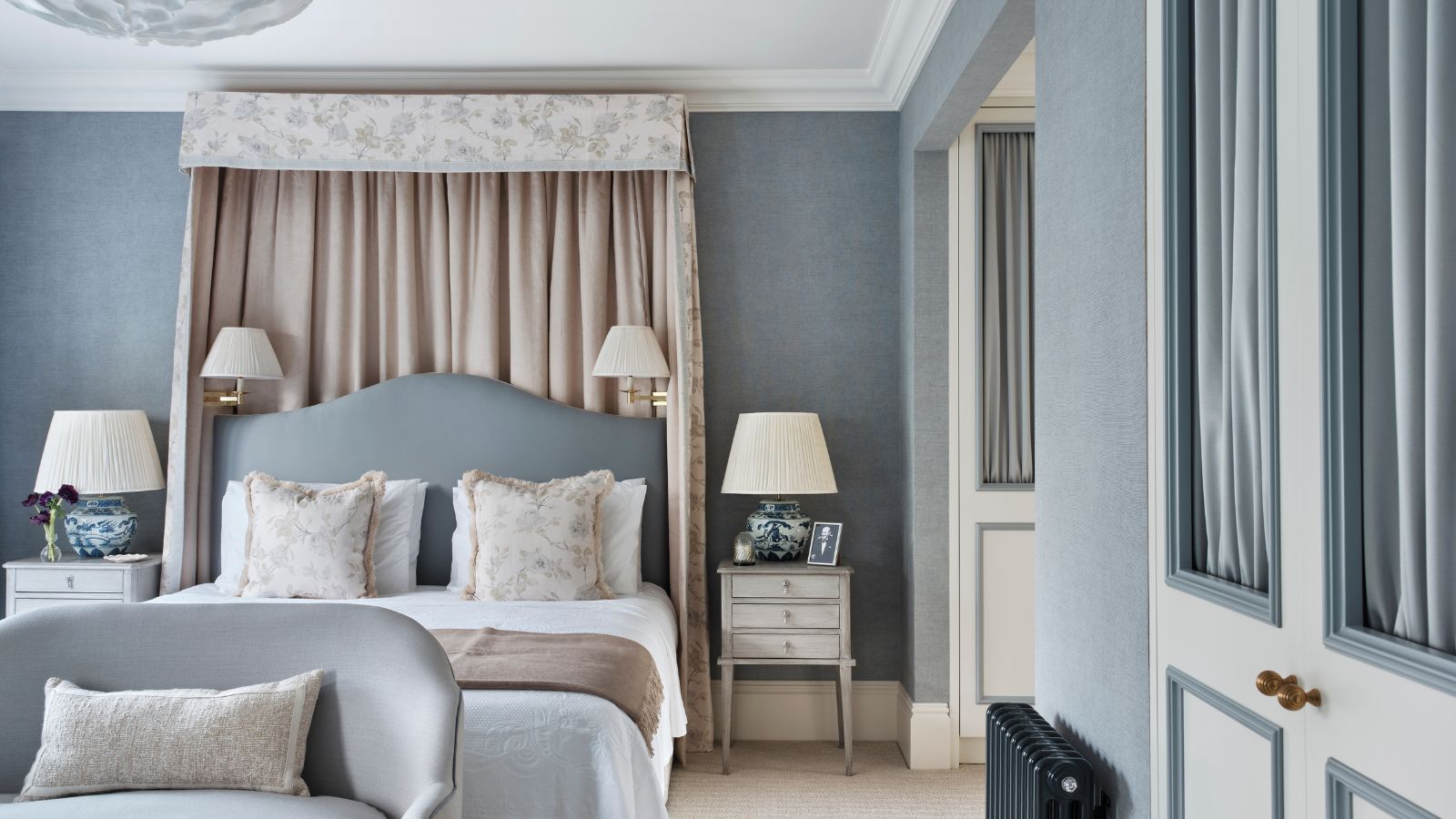 Schlafzimmer mit hellblauen Wänden, einem geblümten Baldachin und einem hellblauen Kopfteil, neutraler Bettwäsche, weißen Vintage-Nachttischen und einem kleinen blauen Zweiersofa