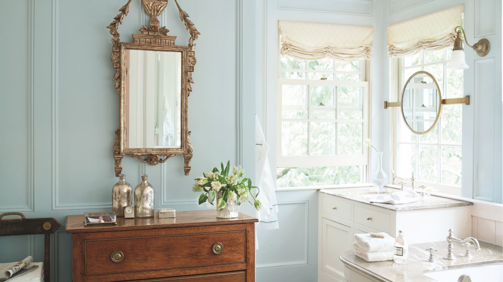 Traditional bathroom with pale blue painted walls, mahogany sideboard with an ornate gold mirror above it, marble bathtub and vanity unit with sink, and two windows with cream roman blinds