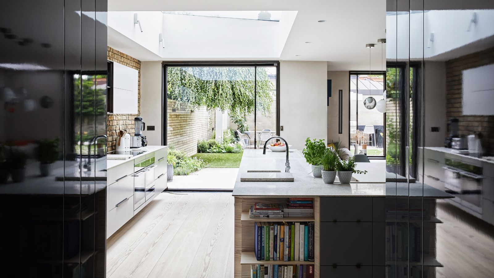 Eine helle und luftige, moderne Küche mit Holzboden, schwarz-weißen Möbeln, einer Insel mit Arbeitsplatten aus weißem Marmor und eingebauten Regalen voller Rezeptbücher, einer unverputzten Backsplash aus Backstein und großen Fenstertüren, die zu einem üppigen grünen Garten führen.