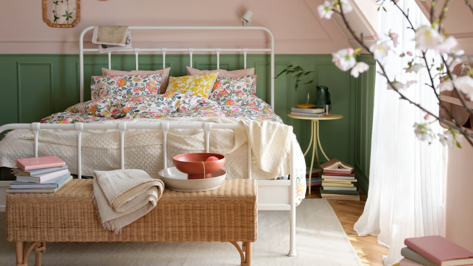 A attic bedroom space with pink and green panelled walls, a large white metal framed double bed with colorful, floral bedding, and a rattan ottoman at the end of it with a stack of books, white blanket, and two stacked bowls in white and orange. Beneath the bed is a white rug and wooden flooring, and to the right is a circular bedside table, stacks of books, large window with sheer white curtains, and a blurred cherry blossom plant in the foreground.