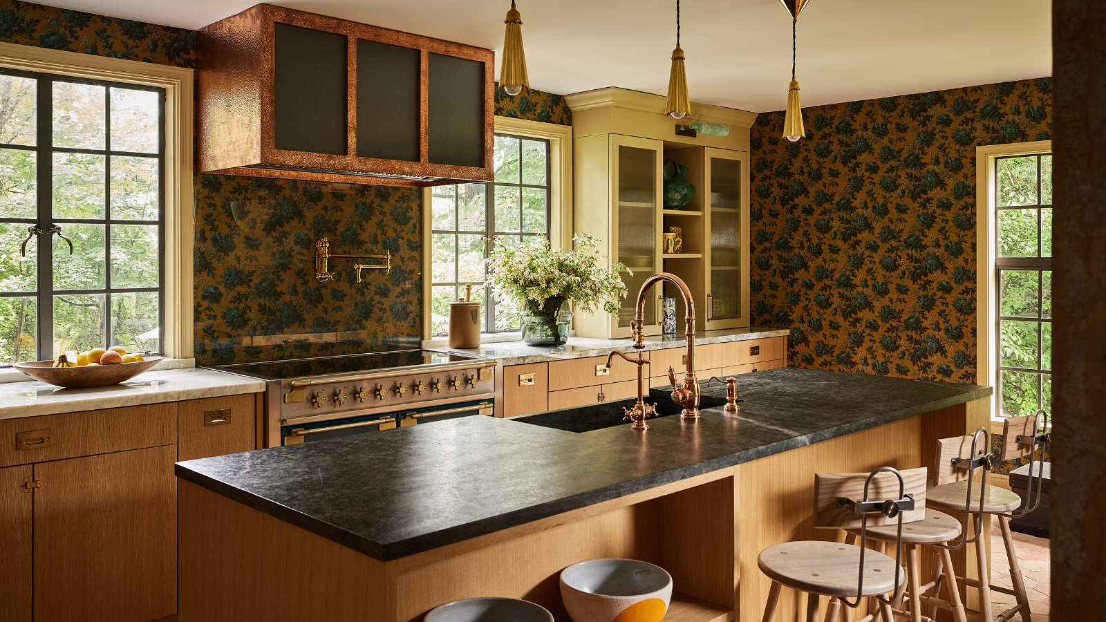 a kitchen with dark floral wallpaper, black stone countertops, a custom copper range hood, and light wood cabinetry