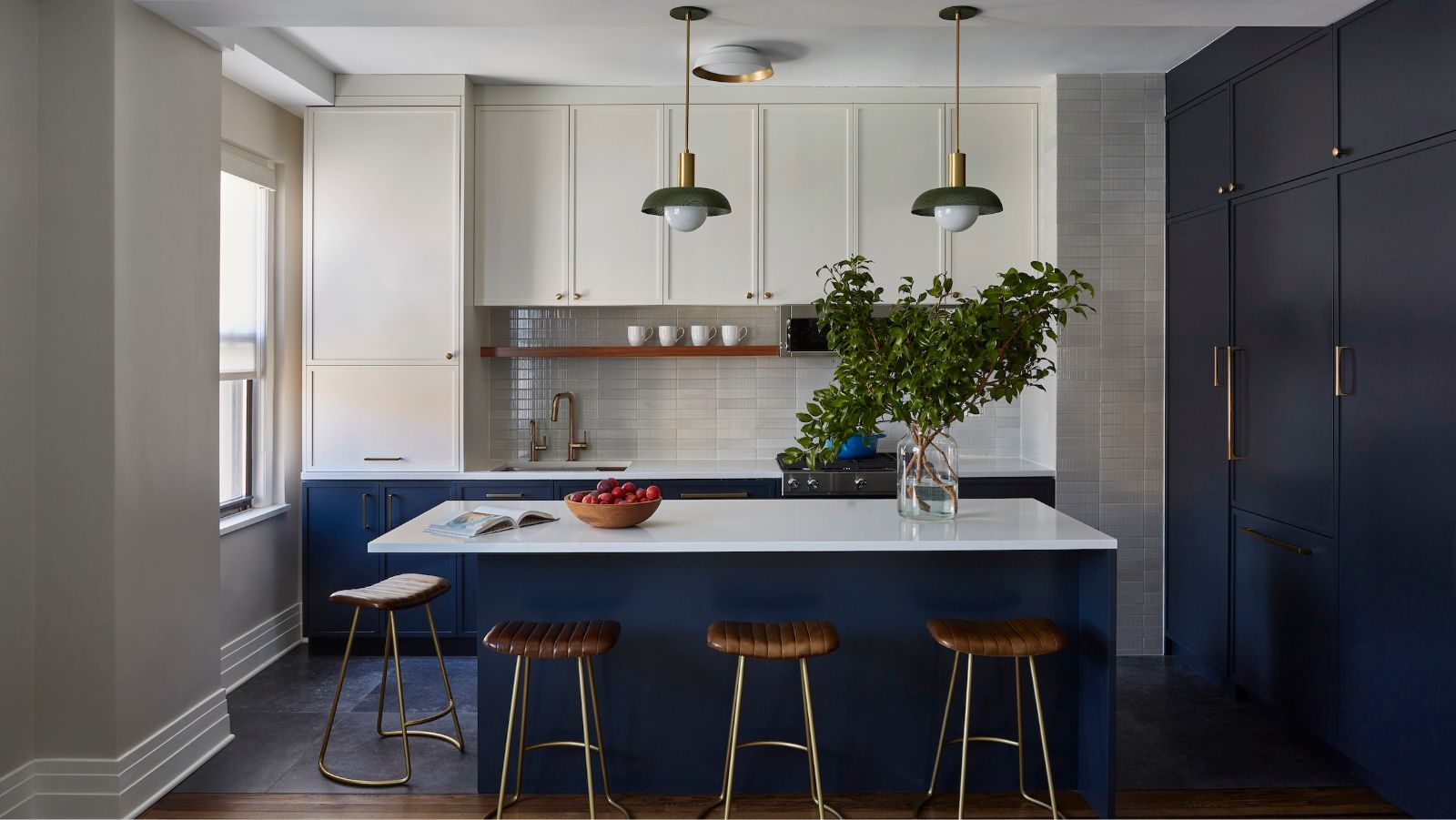 A modern kitchen with dark navy blue cabinets and an island. Gray wall tiles and a row of white upper kitchen cabinets. Brass fixtures on the cabinets, three tan leather bar stools by the island, and a vase of foliage.