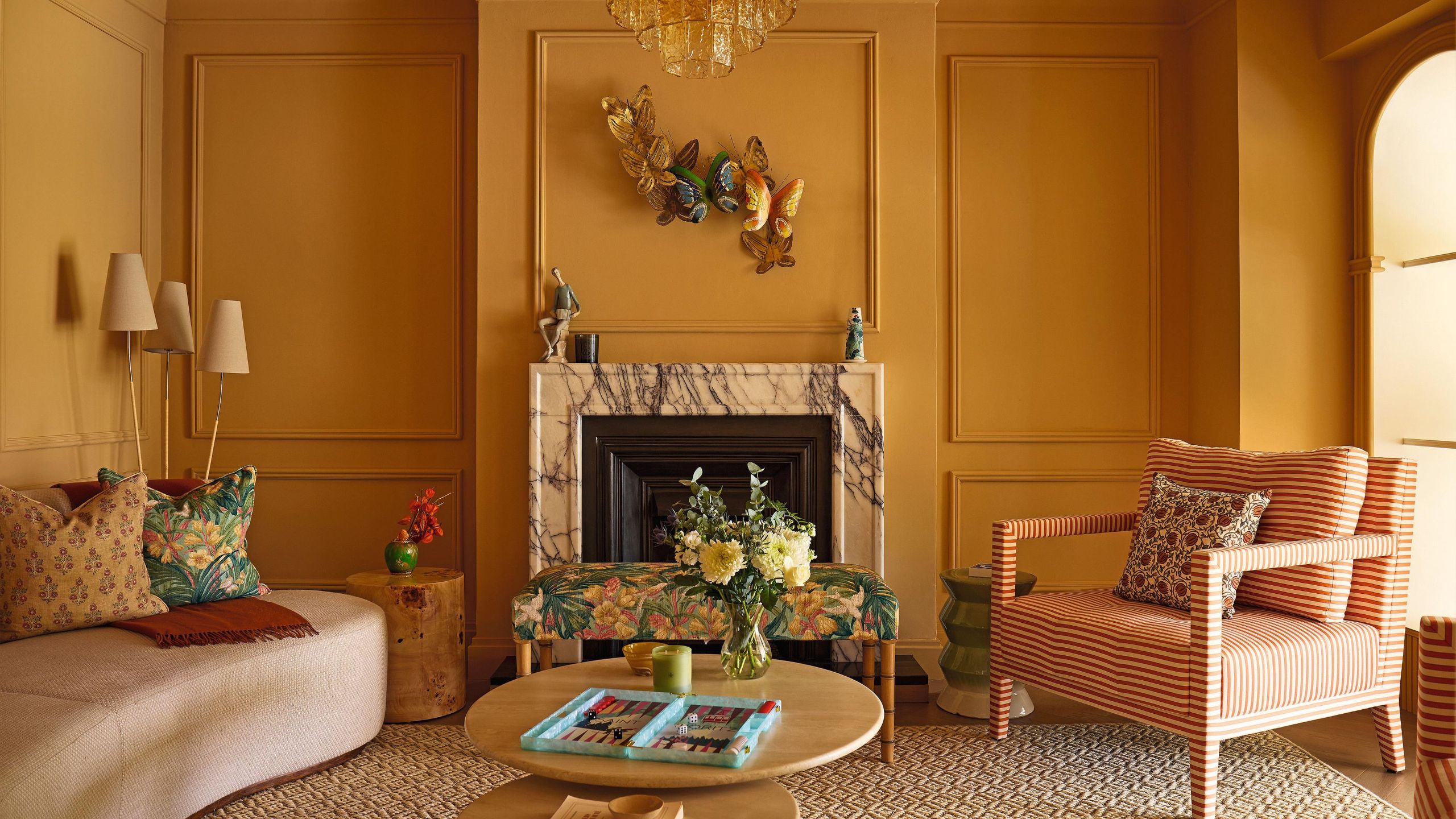 a sunshine yellow living room with wall molding details, a marble fireplace, styled with a curved neutral sofa on the left and a red striped armchair on the right