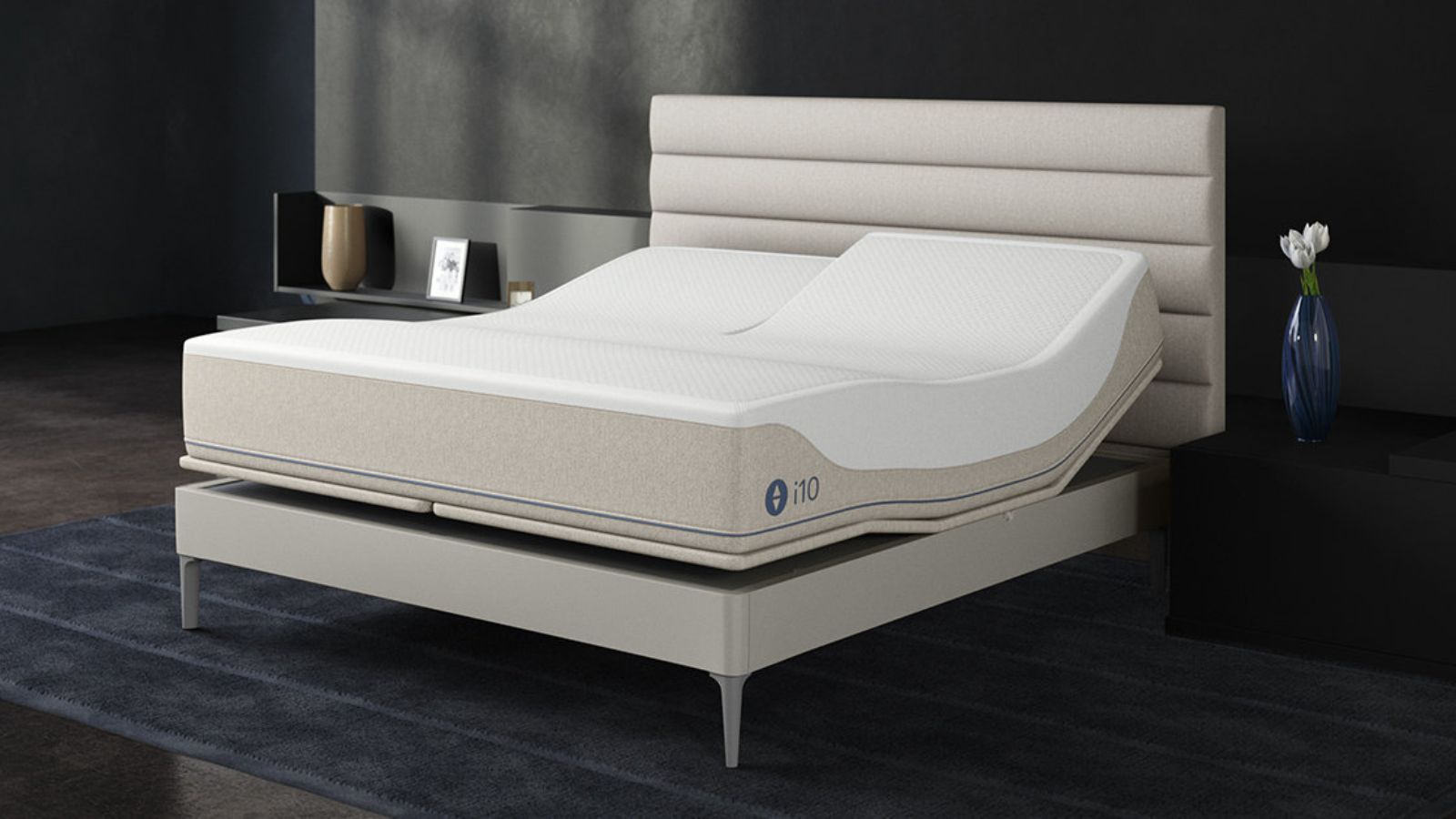 Eine hellbeige Sleep Number-Matratze auf einem Bettgestell mit geriffeltem Kopfteil. Das Bett steht in einem dunklen Raum mit schwarzen Wänden, darunter ein marineblauer Teppich und rechts eine Glasvase voller weißer Blumen. Auf der linken Seite befindet sich ein eingebautes Regal mit goldener Vase und Bilderrahmen.