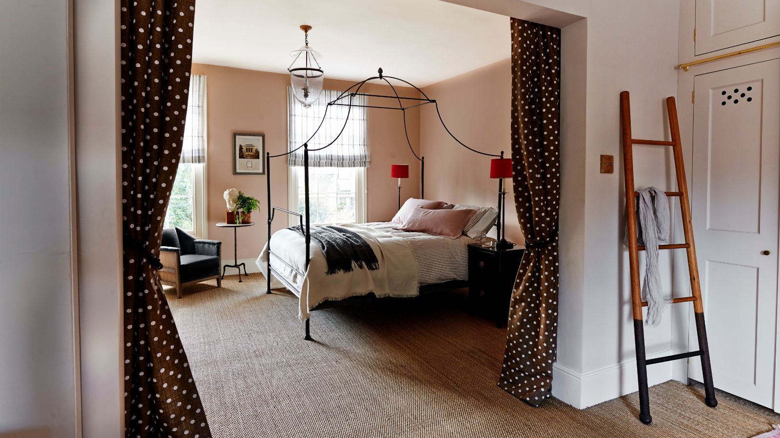 A peach bedroom with a black four-poster bed, polka dot curtains, and a blanket ladder against a linen closet.