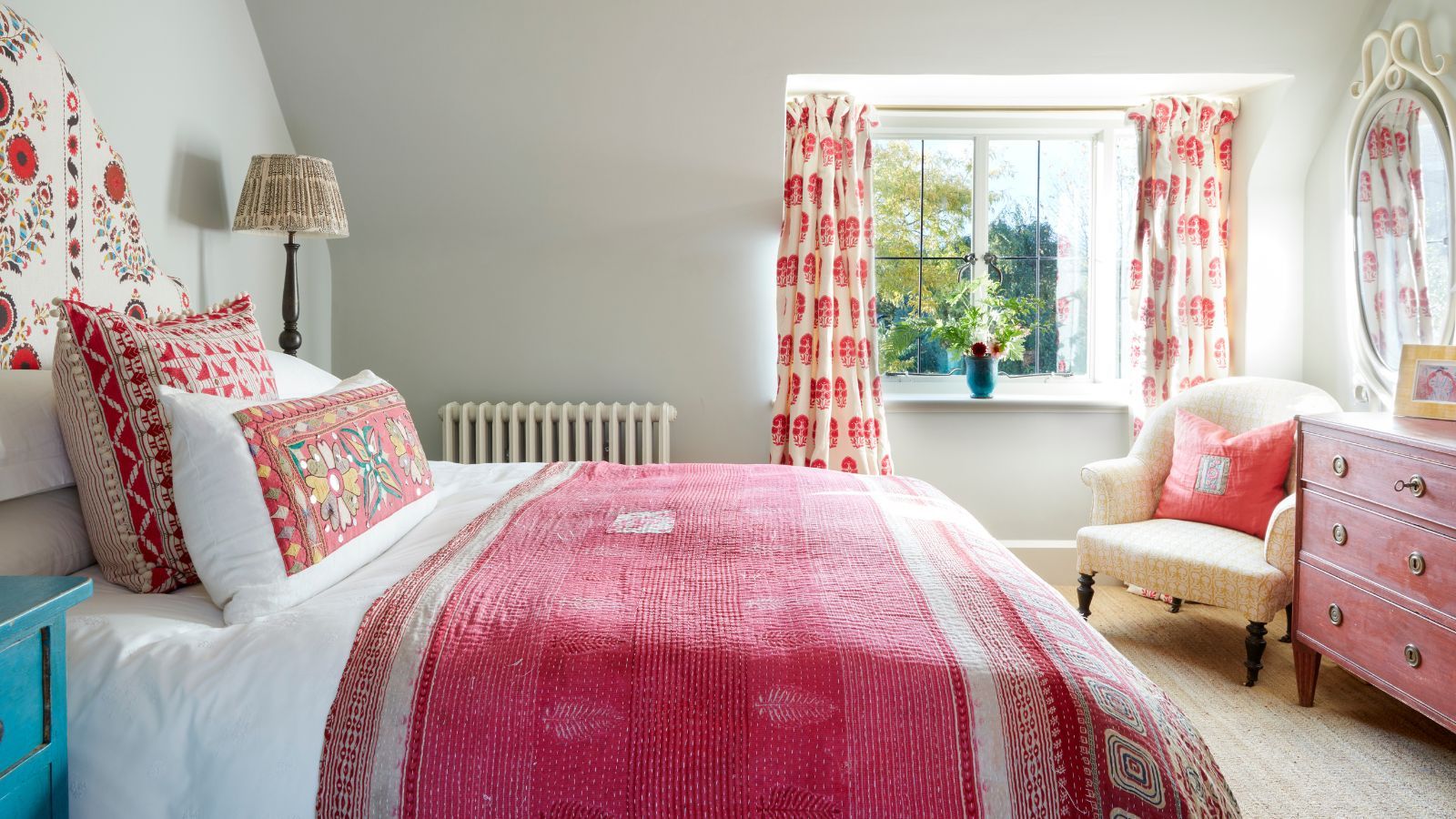 A white bedroom with pink accents with a large double bed with white bedding, pink blankets, pink cushions, and a patterned headboard. In the background is a large window with pink and white curtains, and to the right is a large pink chest of drawers, cream upholstered chair, pink cushion, and mirror.