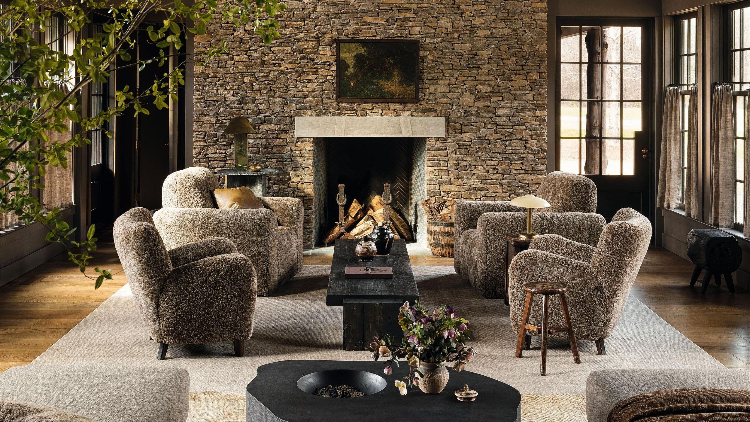 a moody modern farmhouse living room with a large stone fireplace and four sheepskin armchairs styled by the fire on a large rug with dark black wooden rustic tables