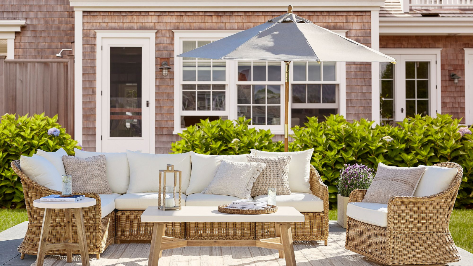 a Cape Cod style outdoor patio with wicker furniture and white cushions, a grey outdoor umbrella, and a cedar shingled house