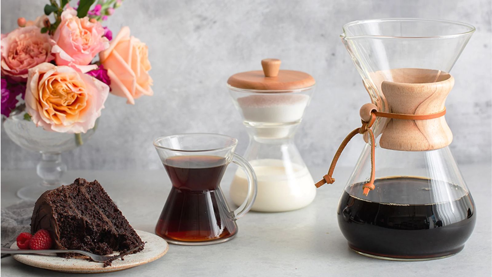 Eine Chemex-Kaffeemaschine, gefüllt mit schwarzem Kaffee, mit einem mit Kaffee gefüllten Glasbecher, einem Milchbehälter aus Glas und einem kleinen Teller Schokoladenkuchen. Dahinter steht eine Glasvase mit Rosen, der Hintergrund ist grau.