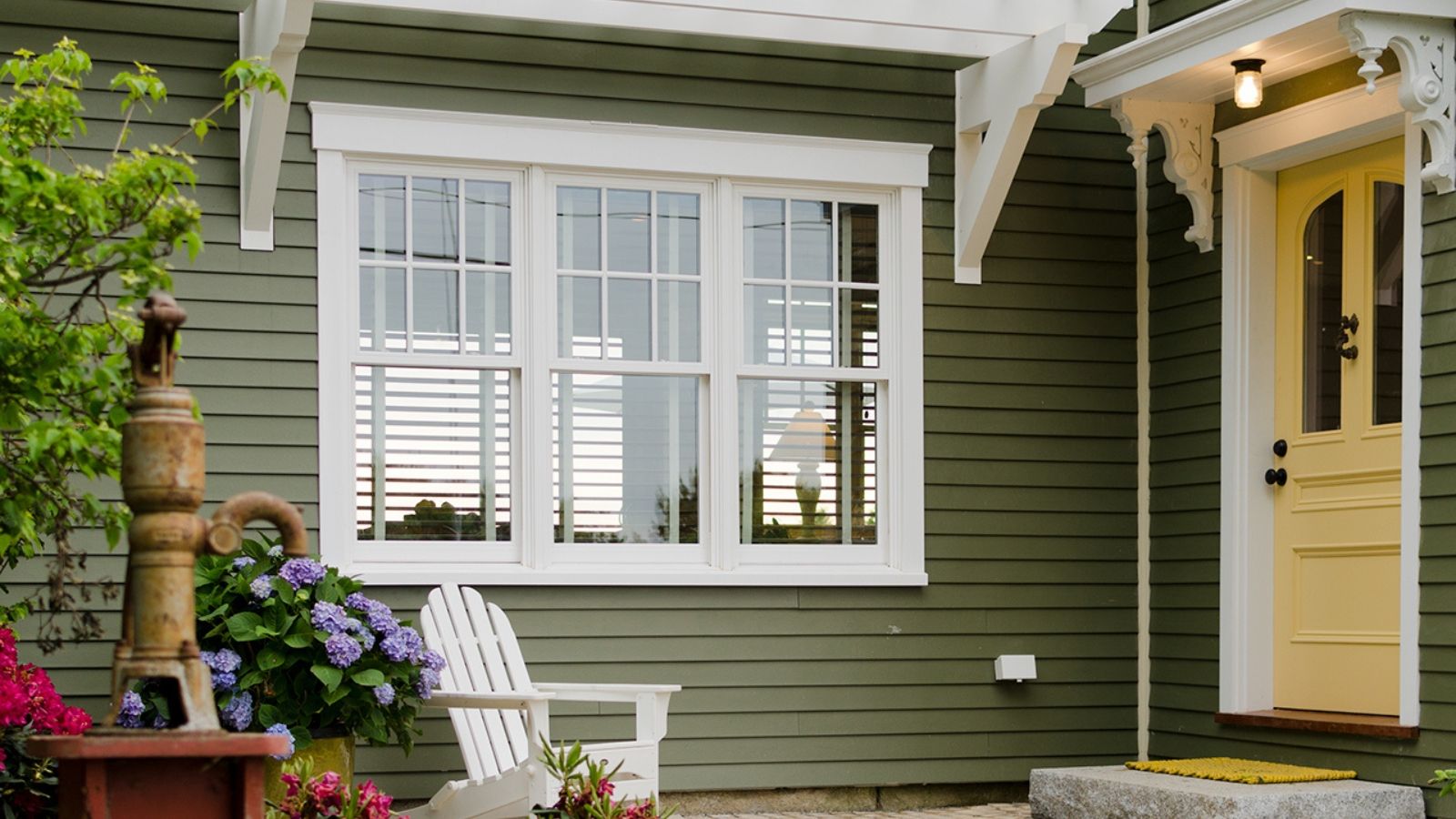 Front porch of a green house with a yellow door. A white chair, a faucet and some flowers are pictured in the foreground.