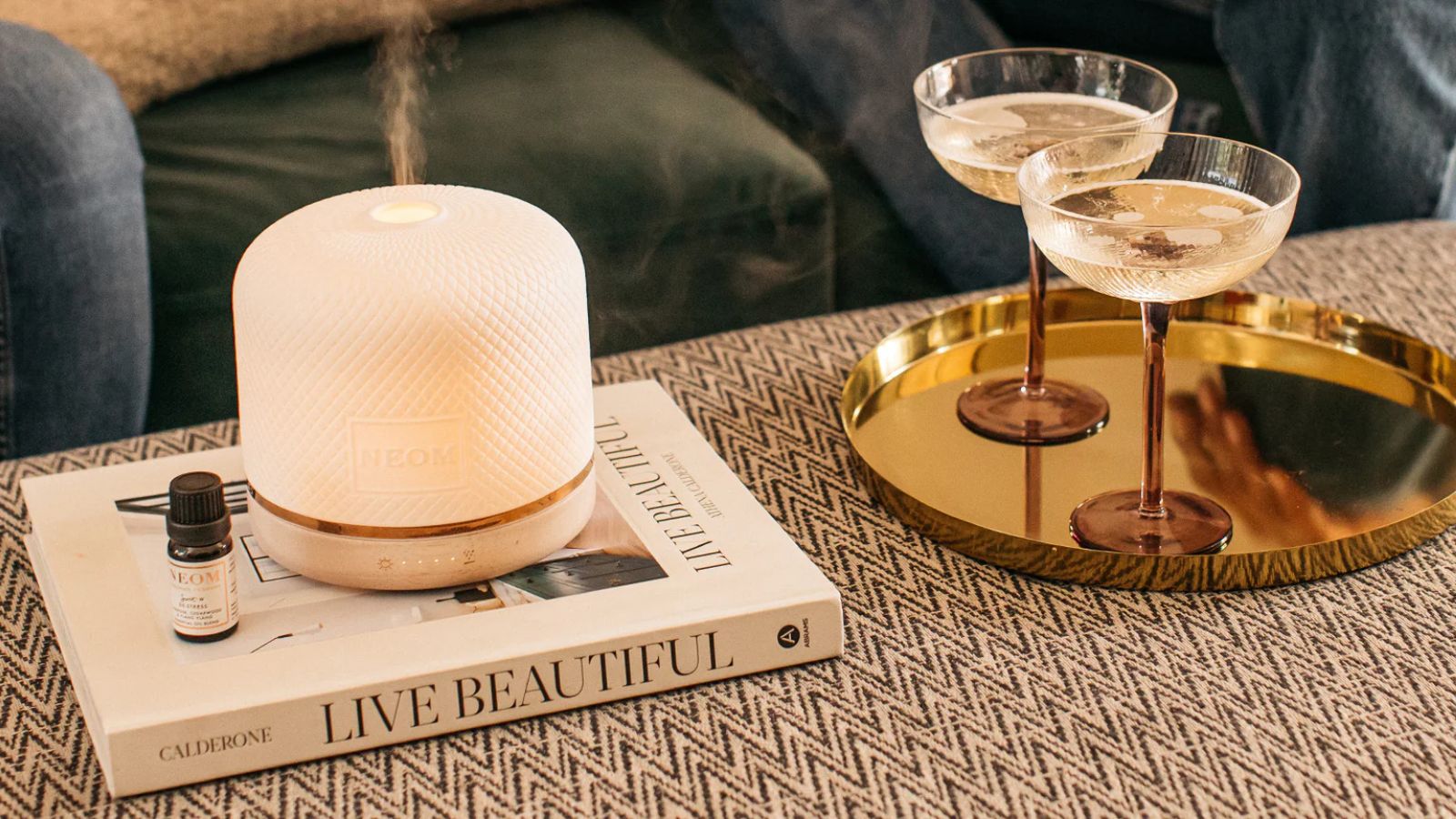 Ein weißer Neom Wellbeing Pod Luxe auf einem weißen Couchtisch und einem gemusterten gepolsterten Ottoman. Rechts ist ein goldenes Tablett mit zwei Champagnerbechern aus braunem und klarem Glas.