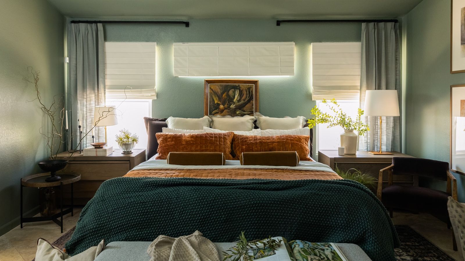Bedroom with teal walls and curtains, white fabric blinds, and a large bed pictured in the center, adorned with multiple blankets, pillows, and cushion. Light is coming through the windows, and there are lamps and potted flowers on the two bedside tables.