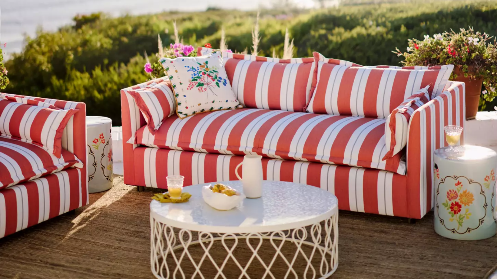 eine rot-weiß gestreifte Outdoor-Couch mit einem Kissen darauf, ein weißer Outdoor-Couchtisch mit dekorativen Details an den Seiten, ein Juteteppich und ein kleiner Akzenttisch rechts von der Couch