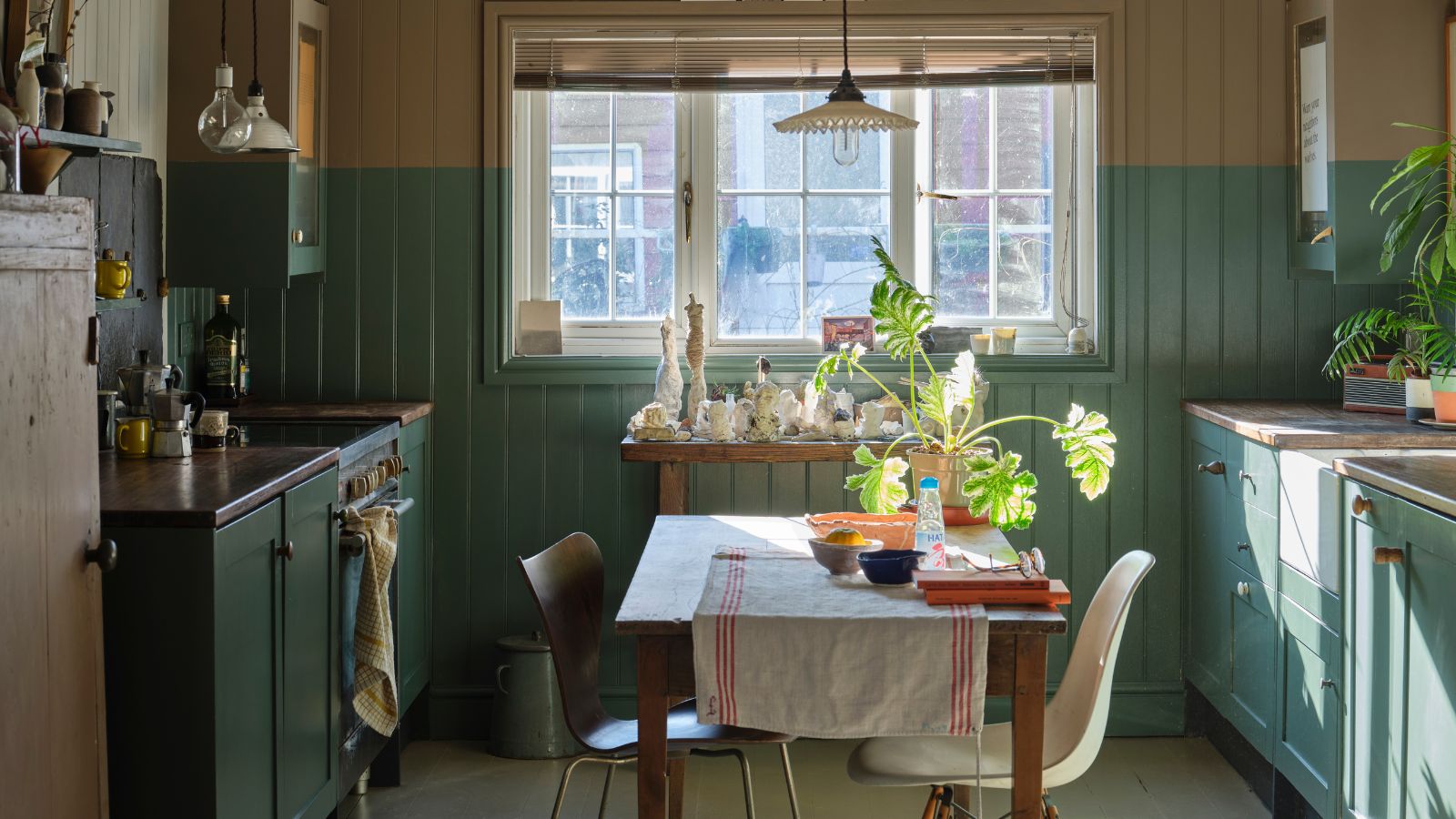 Kleine Küche mit salbeigrünen Schränken links und rechts, mit Fenster in der Mitte hinten und einem Tisch in der Mitte, der Platz mit zwei nicht passenden Stühlen bietet.