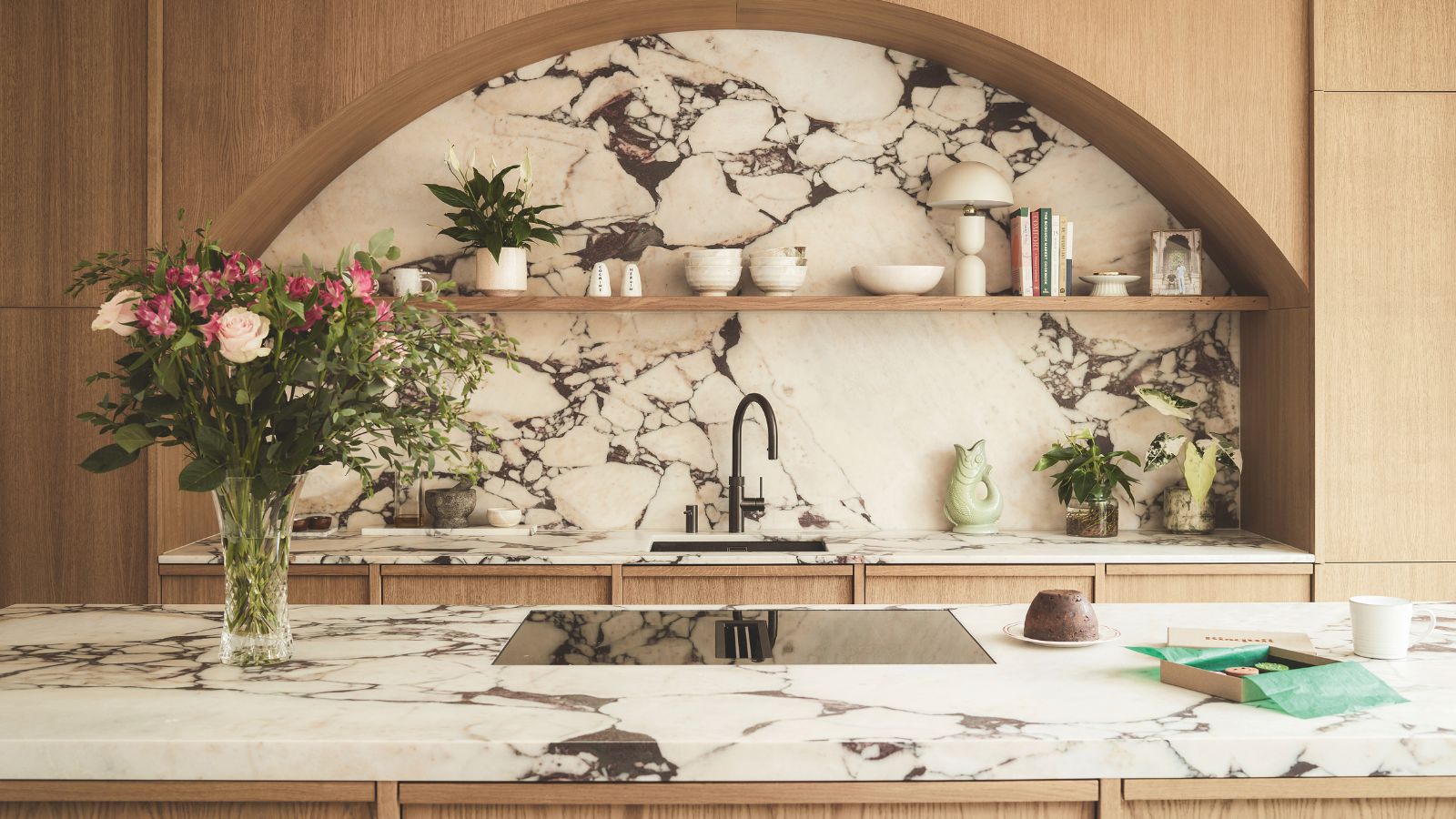 Eine moderne Küche mit gewölbten Armaturen aus hellem Holz über einem Spülbecken und einer großen Marmorrückwand. Eine Insel gegenüber mit passender Marmorplatte und einem Glasherd. Links eine Vase mit Rosen. 