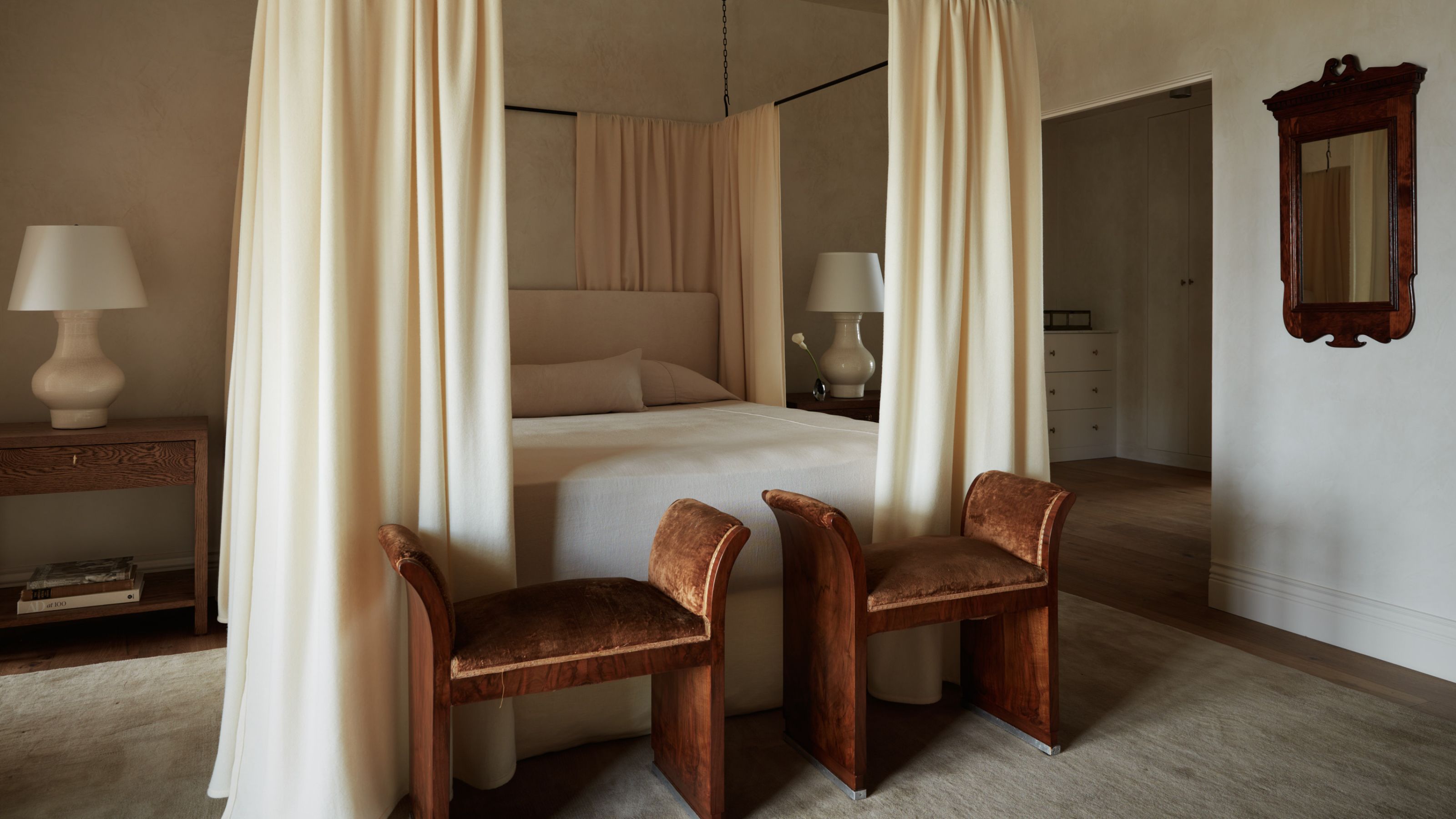 ein ruhiges, neutrales Schlafzimmer mit gekalkten Wänden, einem Himmelbett aus schwarzem Metall mit cremefarbenen Vorhängen, einem neutralen Teppich und zwei schokoladenbraunen Ottomanen am Fußende des Bettes mit antikem Holzspiegel und Nachttisch