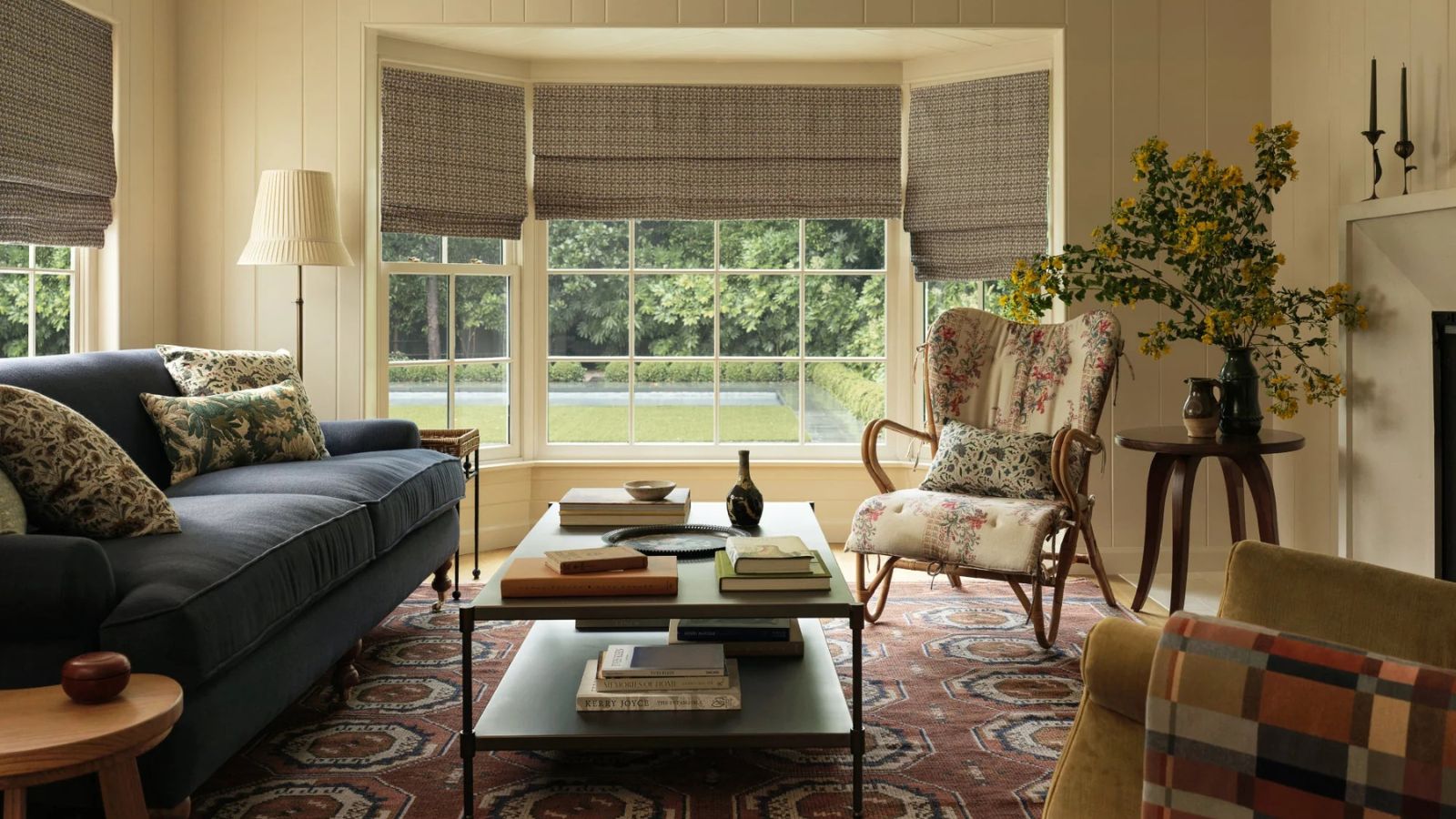 Wohnzimmer mit karierten Jalousien, großem roten und blauen Teppich, samtblauem Sofa, geblümtem Sessel und Couchtisch mit Büchern und Vase